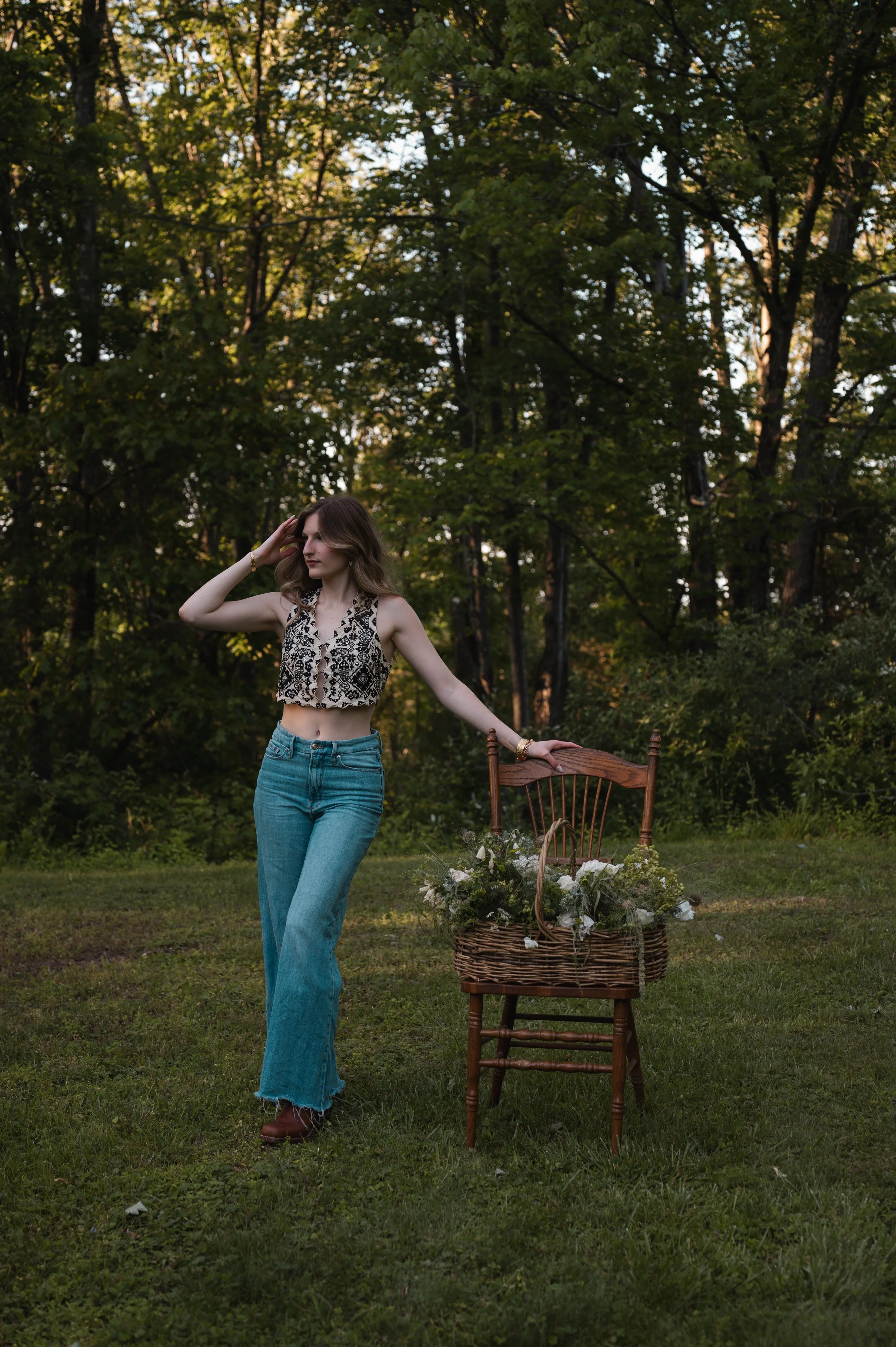 A woman with wavy brown hair wearing a black and white patterned crop top, blue jeans, and brown boots standing outdoors in a grassy area surrounded by trees. She is touching her head with her right hand and has her left hand resting on a wooden chair next to a wicker basket filled with white flowers.