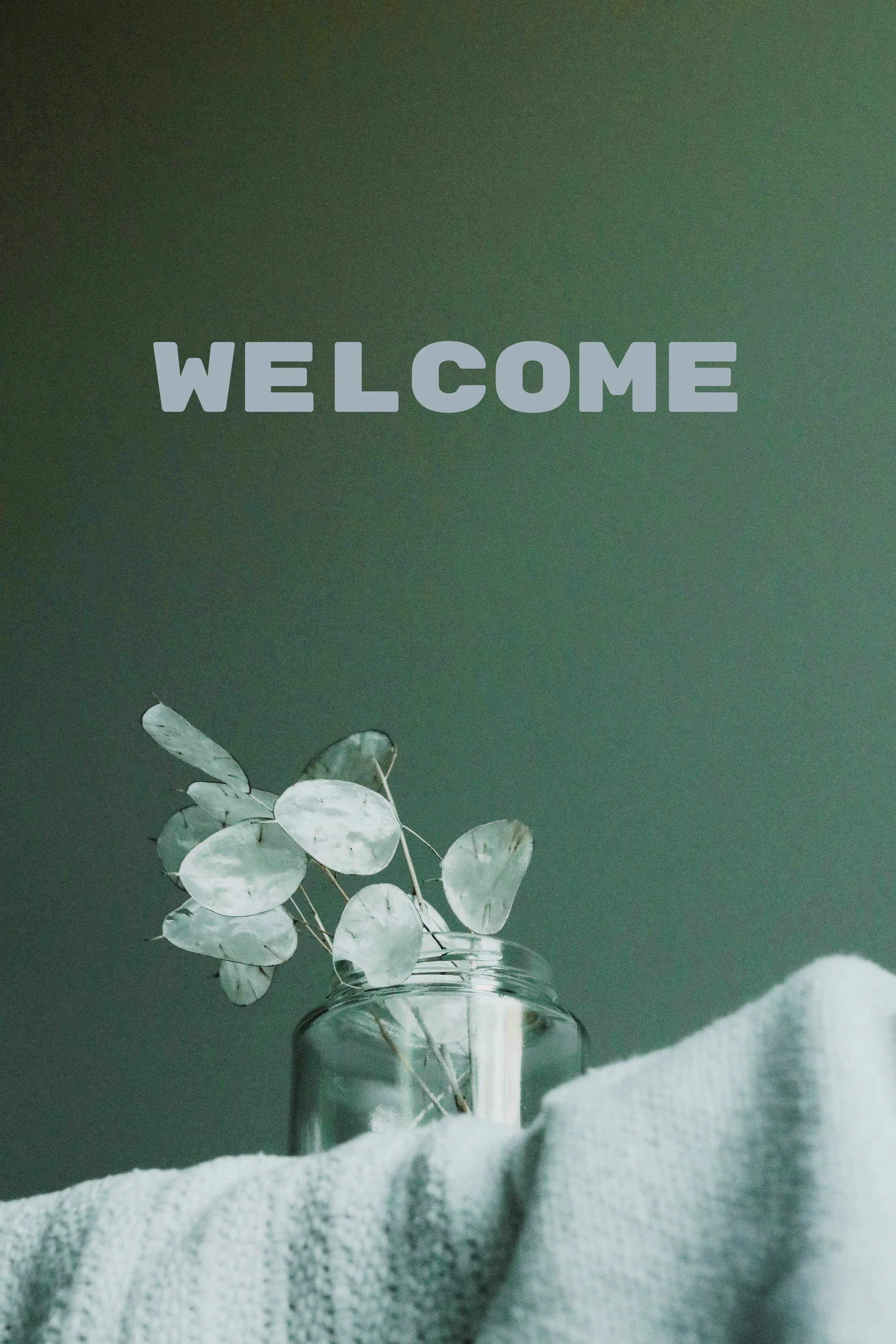 A vase of dried eucalyptus leaves on a table with a textured cloth, against a dark green background with the word "WELCOME" in large gray letters.