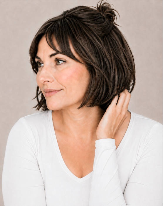 A woman with shoulder-length brown hair and blue eyes, wearing a white long-sleeve shirt, is touching her neck and looking to her left against a gray background.
