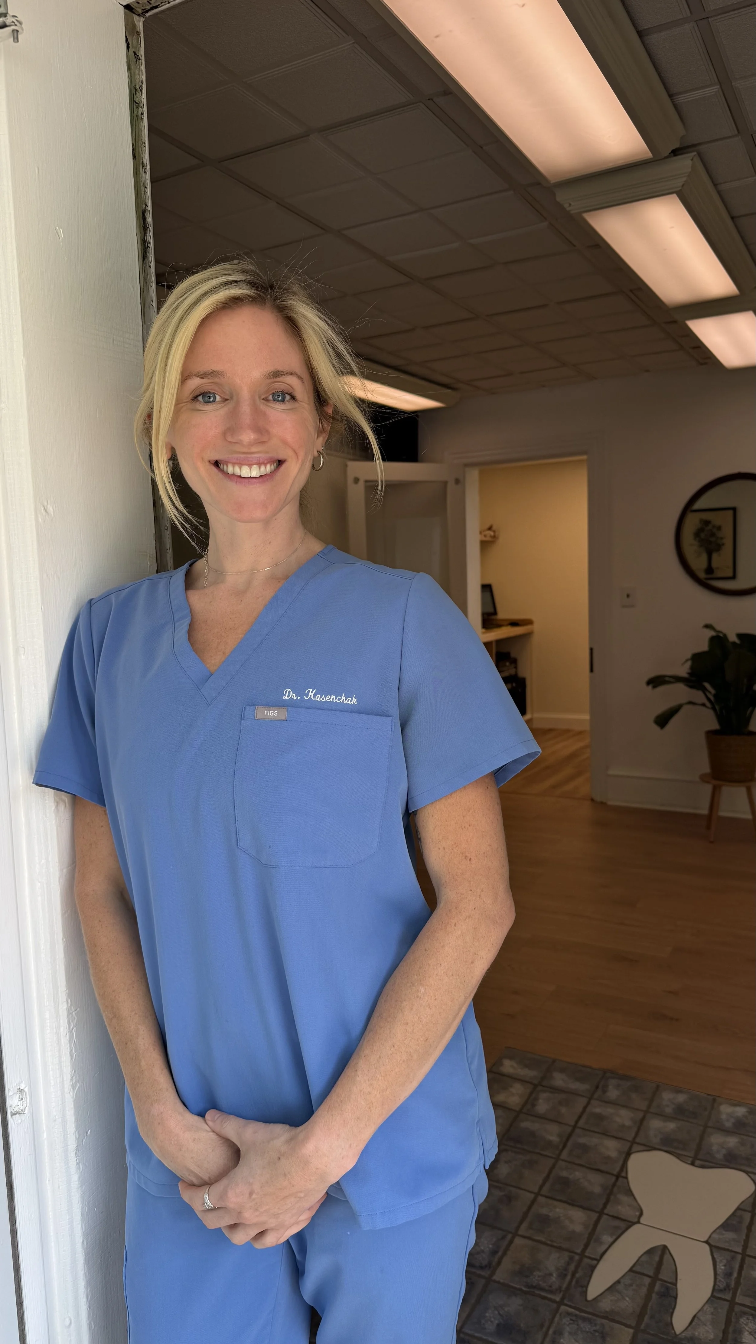 A woman in blue medical scrubs standing in a room with warm lighting, wooden flooring, a potted plant, and wall decor including a framed picture of a tree.