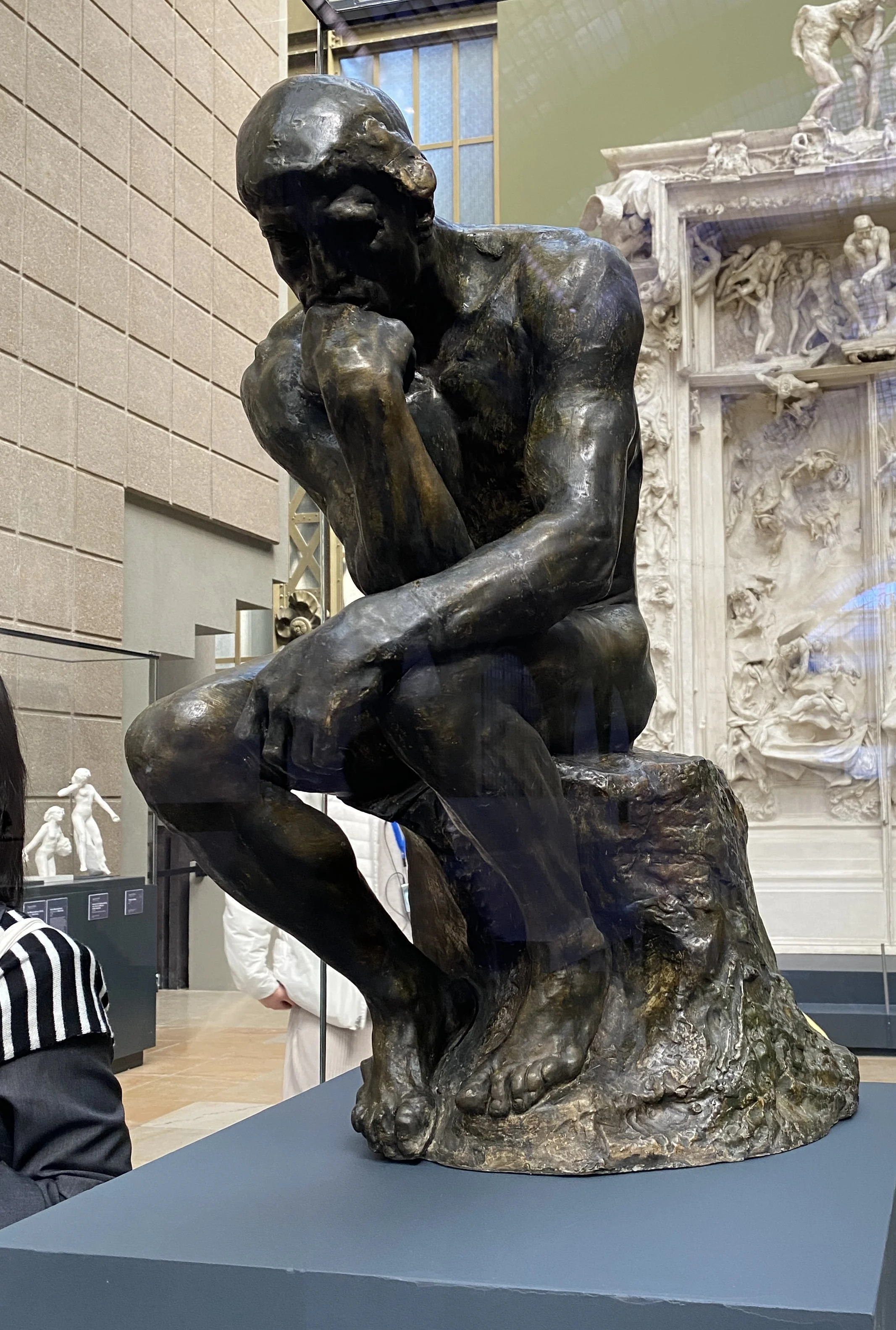 Bronze sculpture of a contemplative man resting his chin on his hand, seated on a rock, displayed on a blue pedestal in a museum setting.