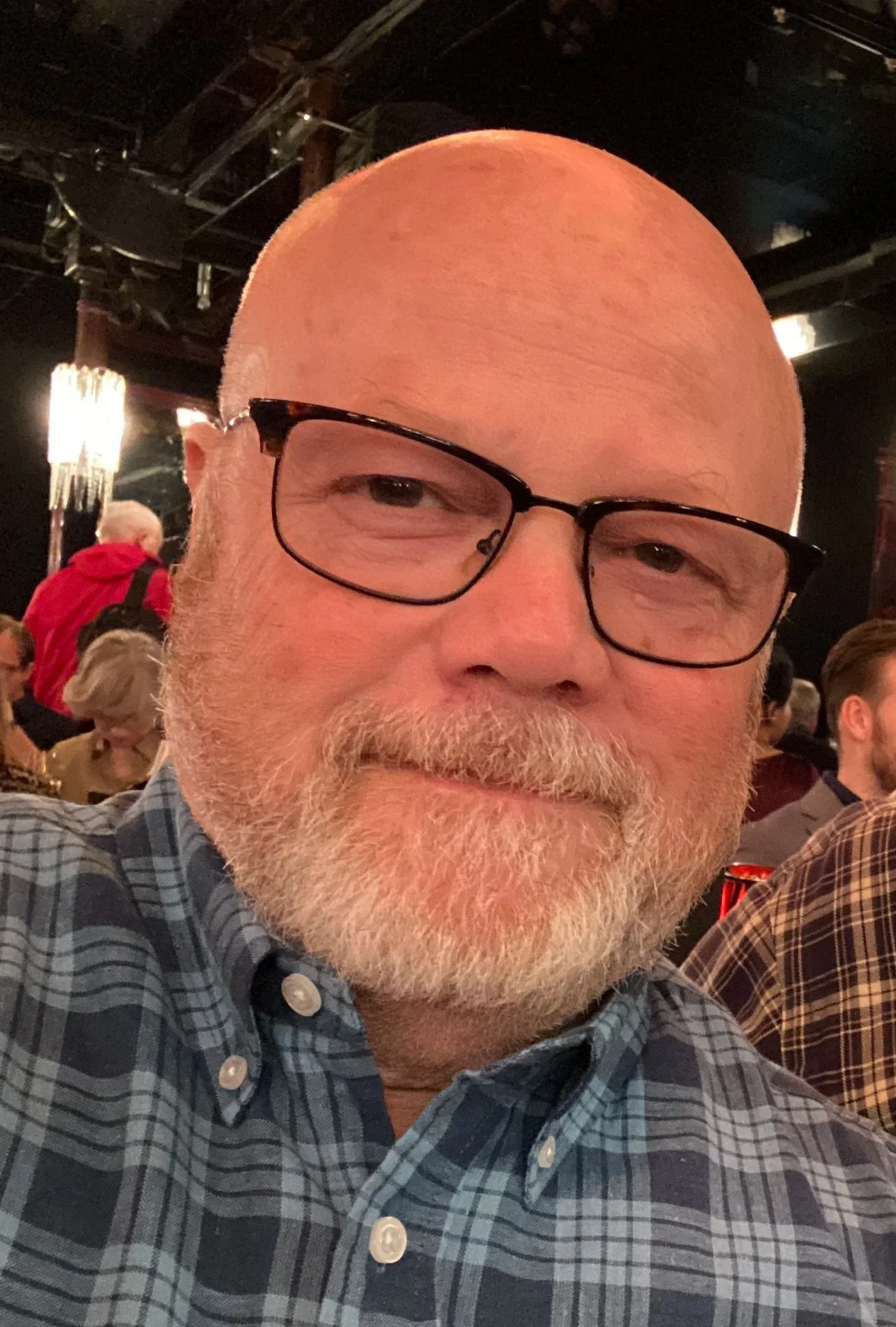 Close-up of a bald man with glasses and a white beard, wearing a plaid shirt, at a social event or gathering.