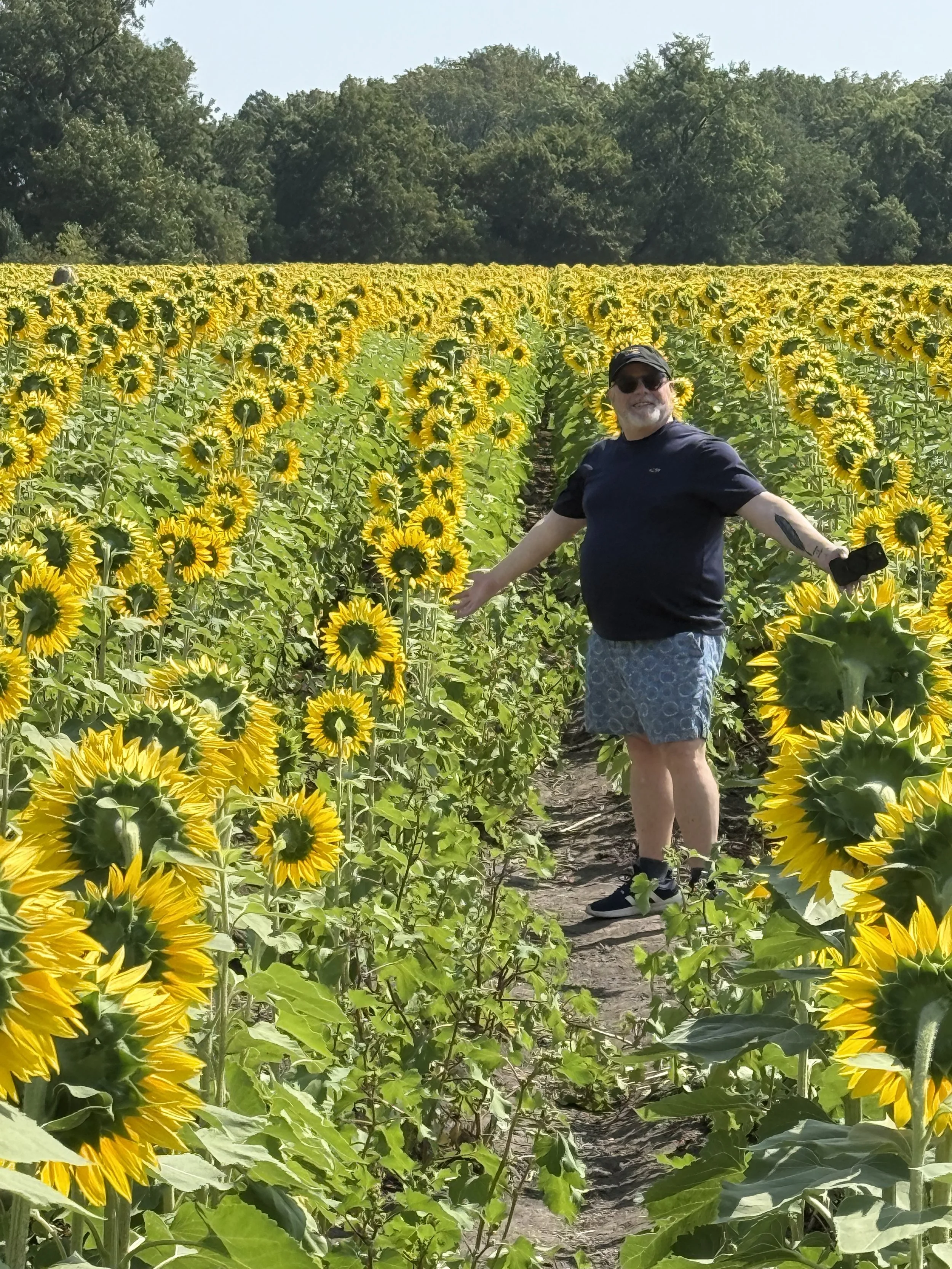 A man wearing sunglasses, a black shirt, and shorts standing in the middle of a sunflower field with arms outstretched, smiling. The sunflower field is lush and filled with tall, bright yellow sunflowers under a clear sky, with trees in the background.