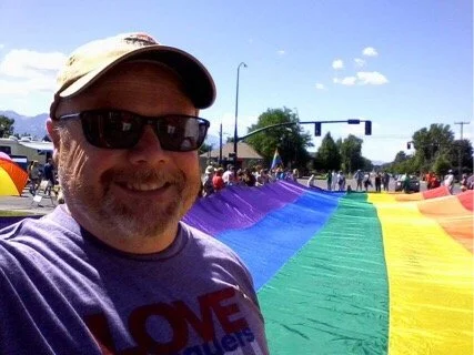 A smiling man wearing sunglasses and a baseball cap takes a selfie outdoors at a pride event with a rainbow flag extending behind him.