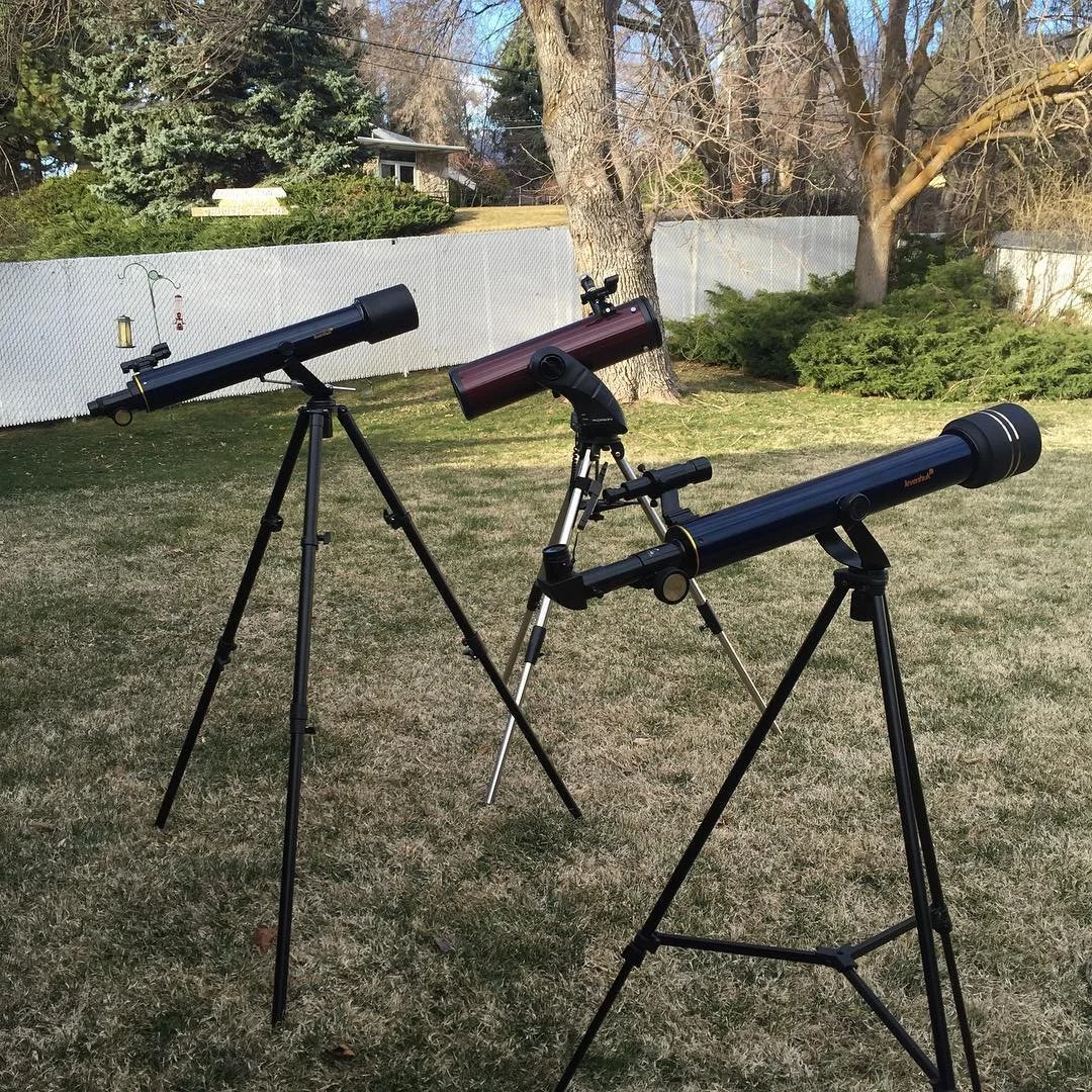 Three telescopes of different sizes set up in a backyard with grass, trees, and a white fence in the background.