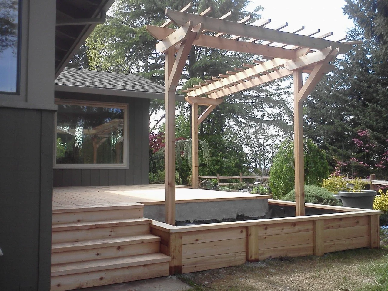 Photo of a house with a wooden deck and a garden with trees and bushes. A new wooden pergola frame is being built over a small raised garden bed next to the deck.
