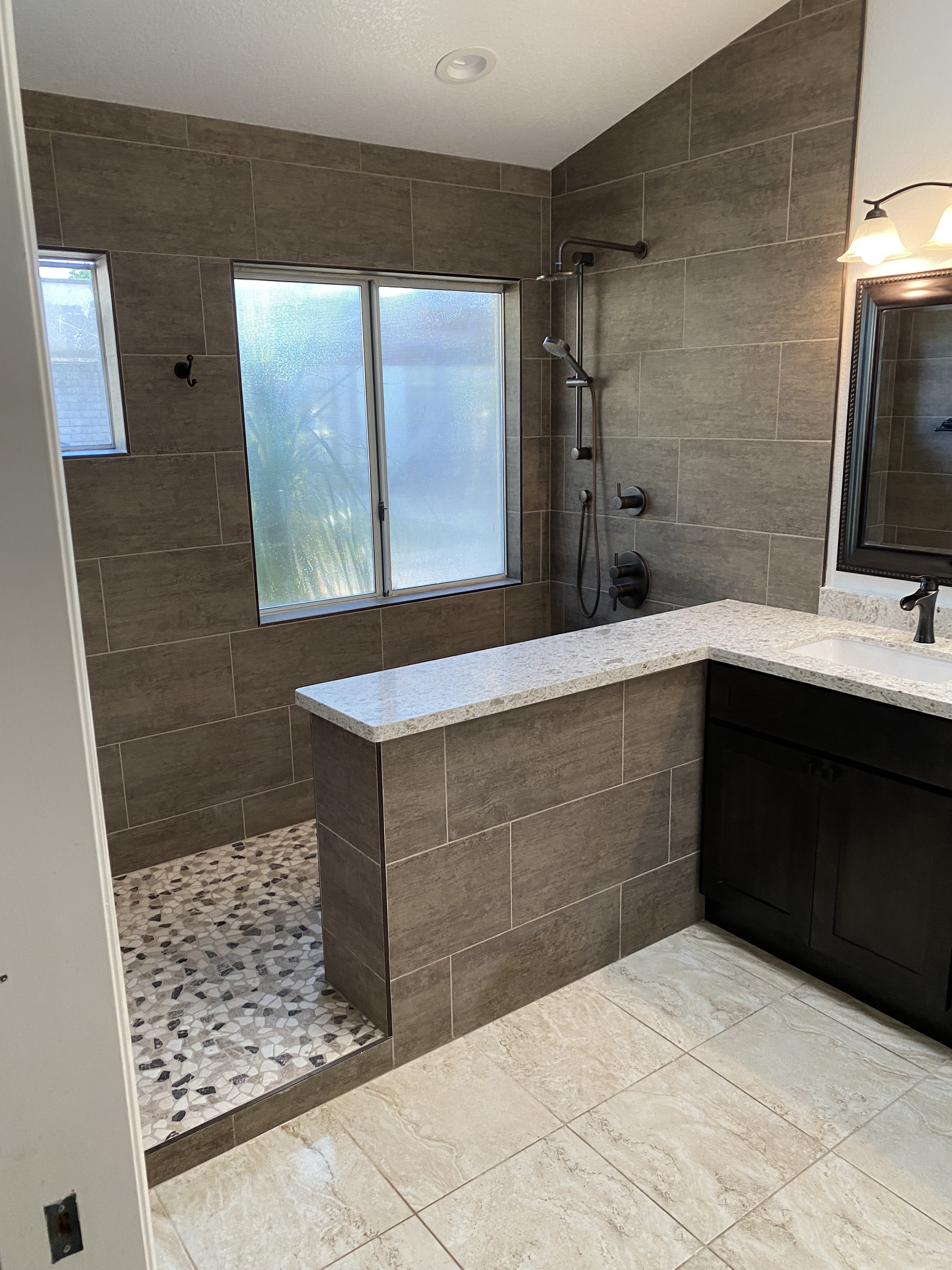 A modern bathroom with a walk-in shower with beige and gray tiles, a handheld shower head, a small window, a vanity with a black cabinet and a white countertop, a mirror with a black frame, and a light fixture with two lamps.