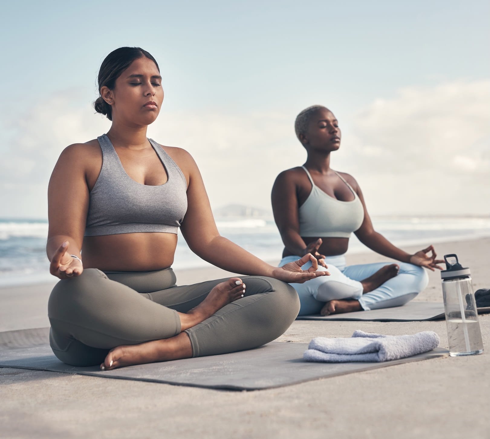 Two women practicing meditation on a beach, sitting cross-legged in a peaceful pose with eyes closed, wearing athletic wear, with a water bottle and towel nearby.