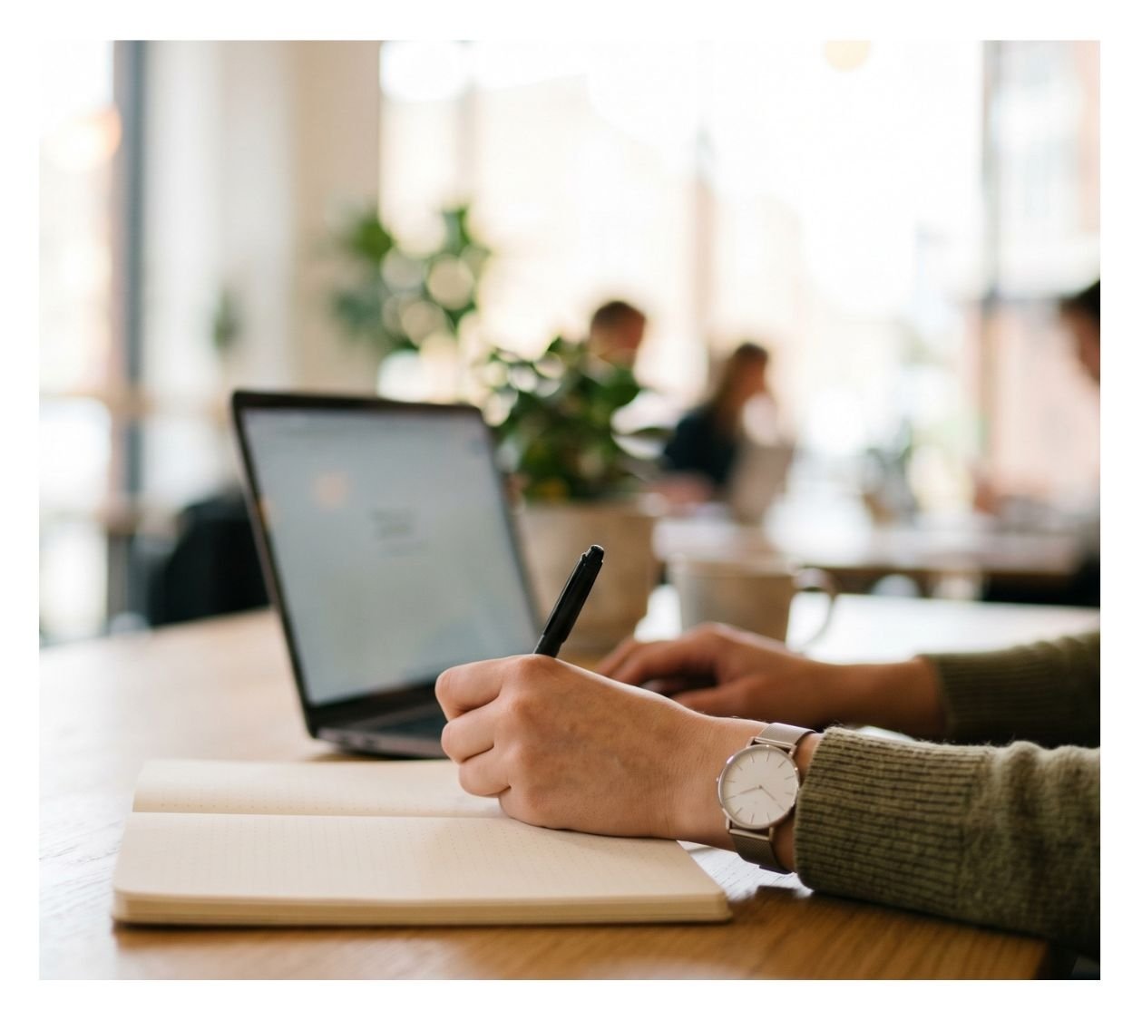 Person writing in notebook with a pen at a desk, with a laptop, in a bright, modern office or cafe environment.