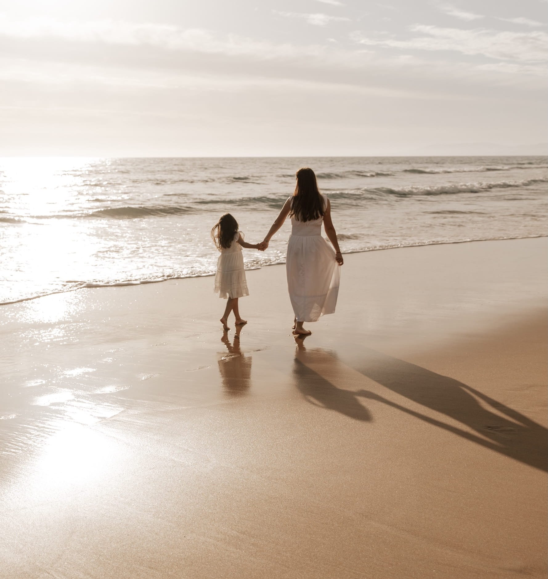 A woman and a young girl walk hand in hand along a sandy beach near the ocean, with gentle waves and a cloudy sky in the background. Energy Healing.