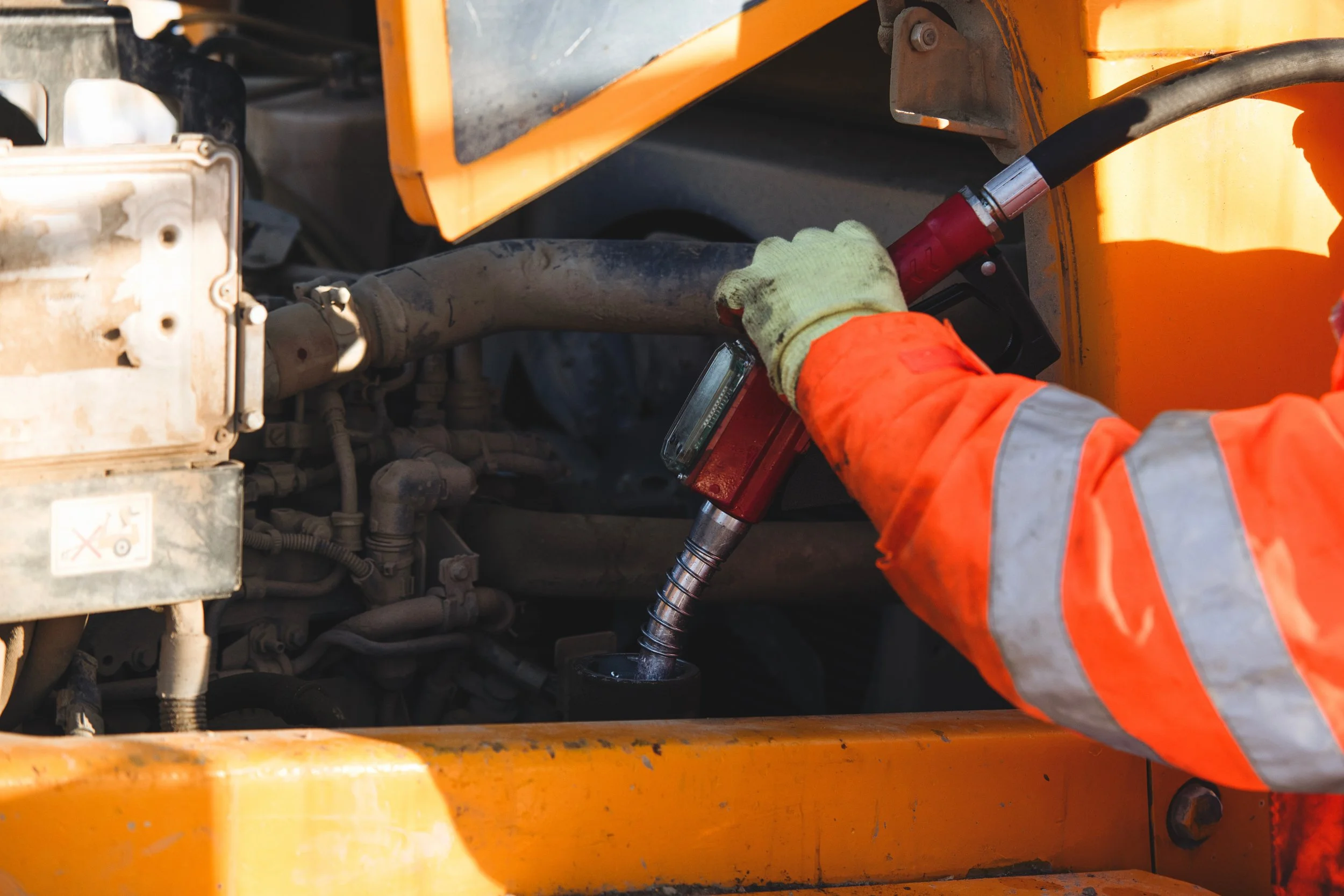 Close-up of a worker in an orange safety jacket and gloves using a hydraulic tool on machinery inside a yellow construction vehicle or equipment.
