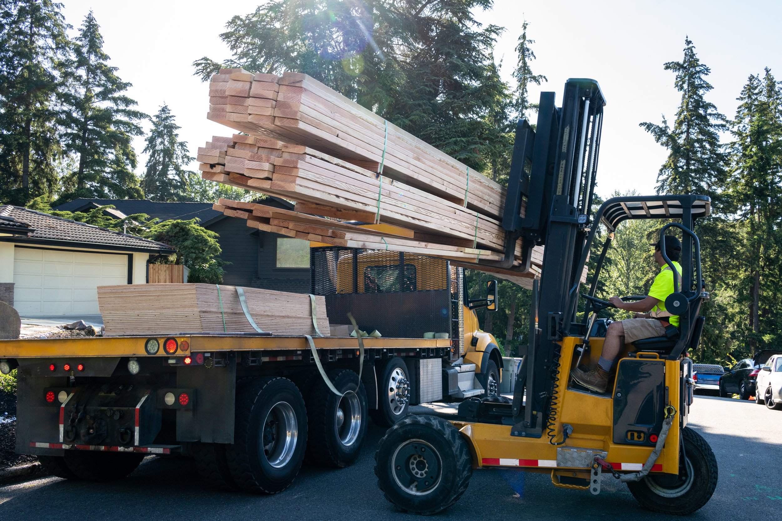 A construction worker operating a forklift truck lifting long wooden planks onto a flatbed truck in a residential neighborhood with houses and trees in the background.