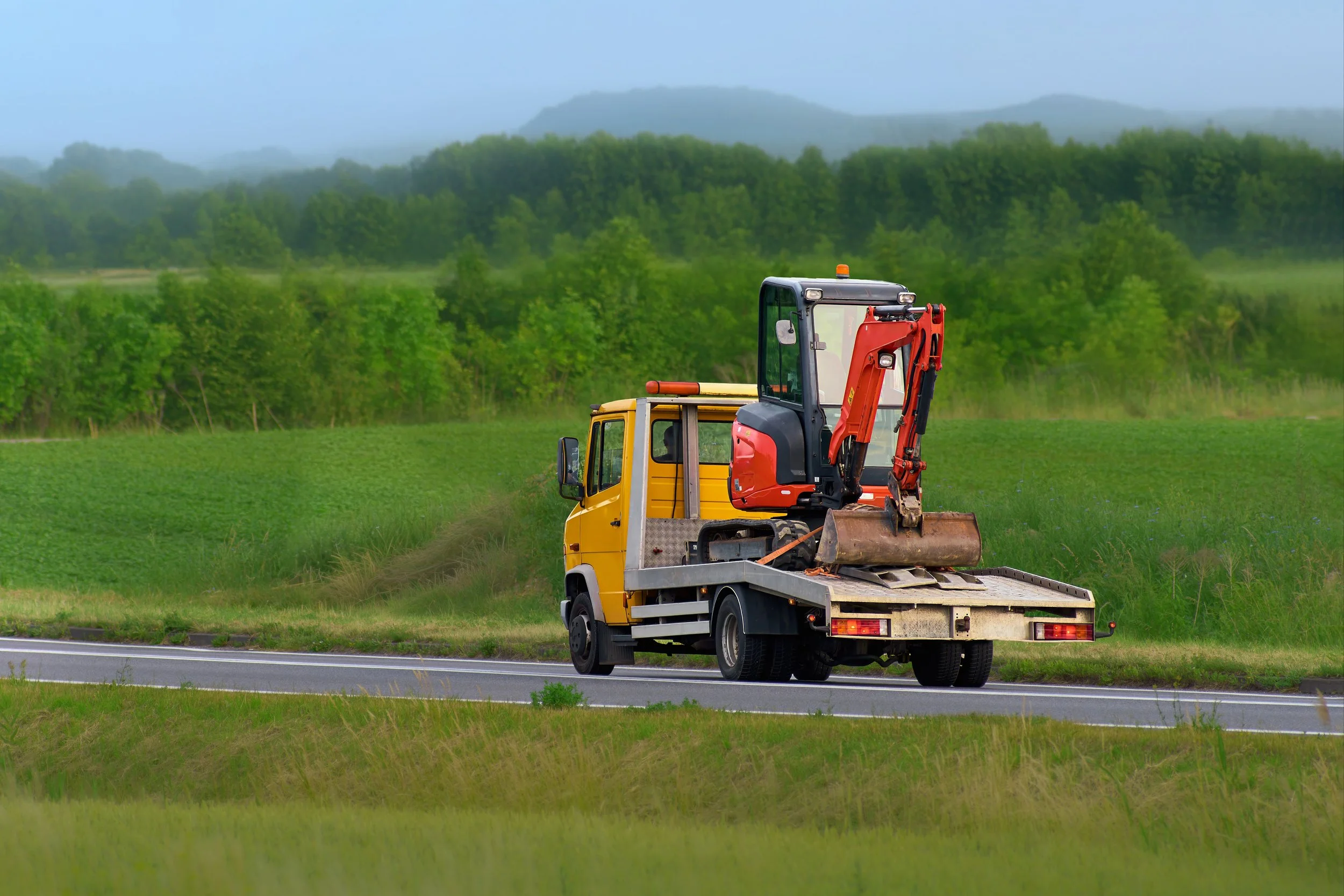 A yellow flatbed truck hauling a red and black mini excavator on a grassy roadside with green trees and mountains in the background.