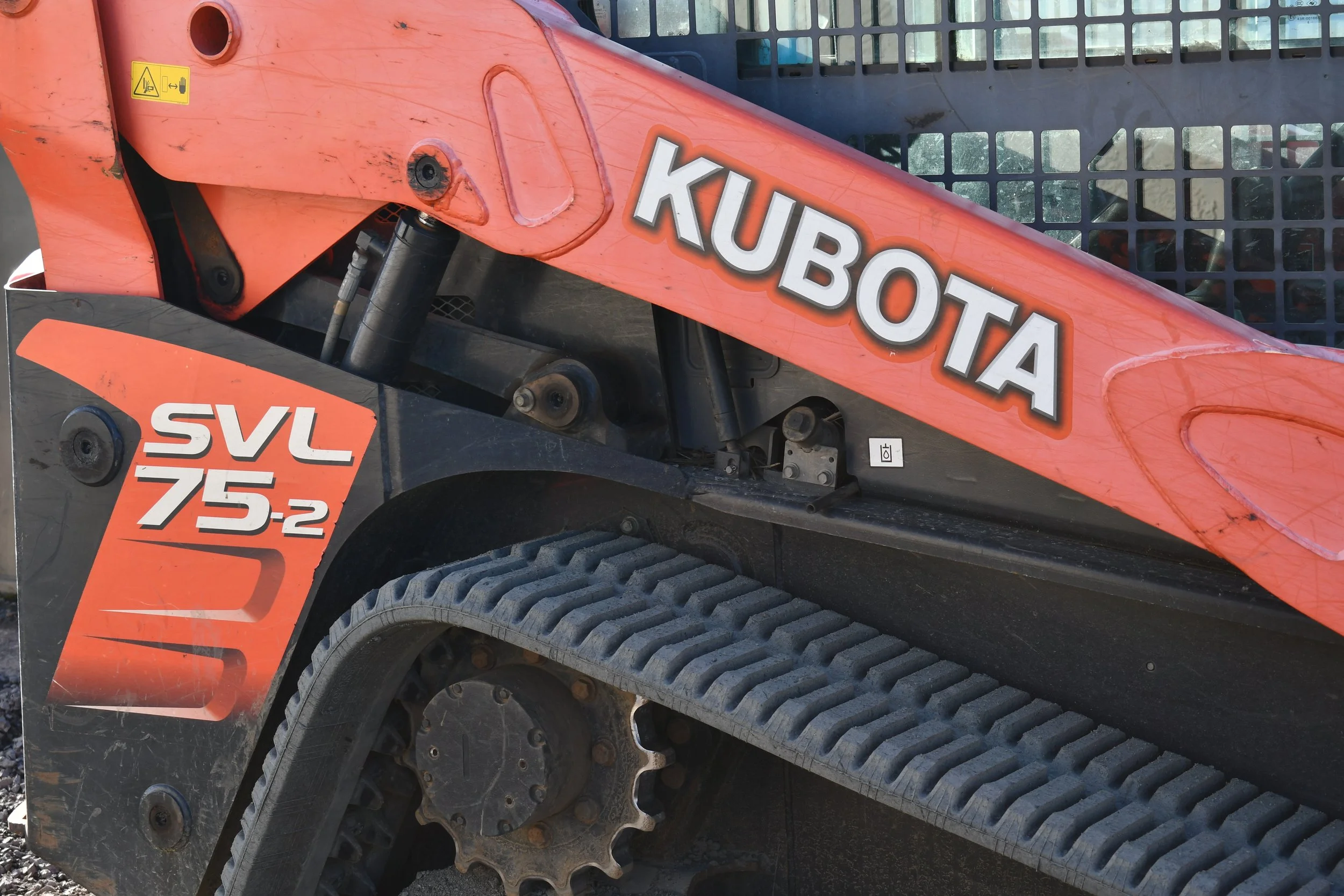 Close-up of an orange Kubota construction machine, showing part of the arm, hydraulic components, and tracks, with black and orange decals and a warning sticker.