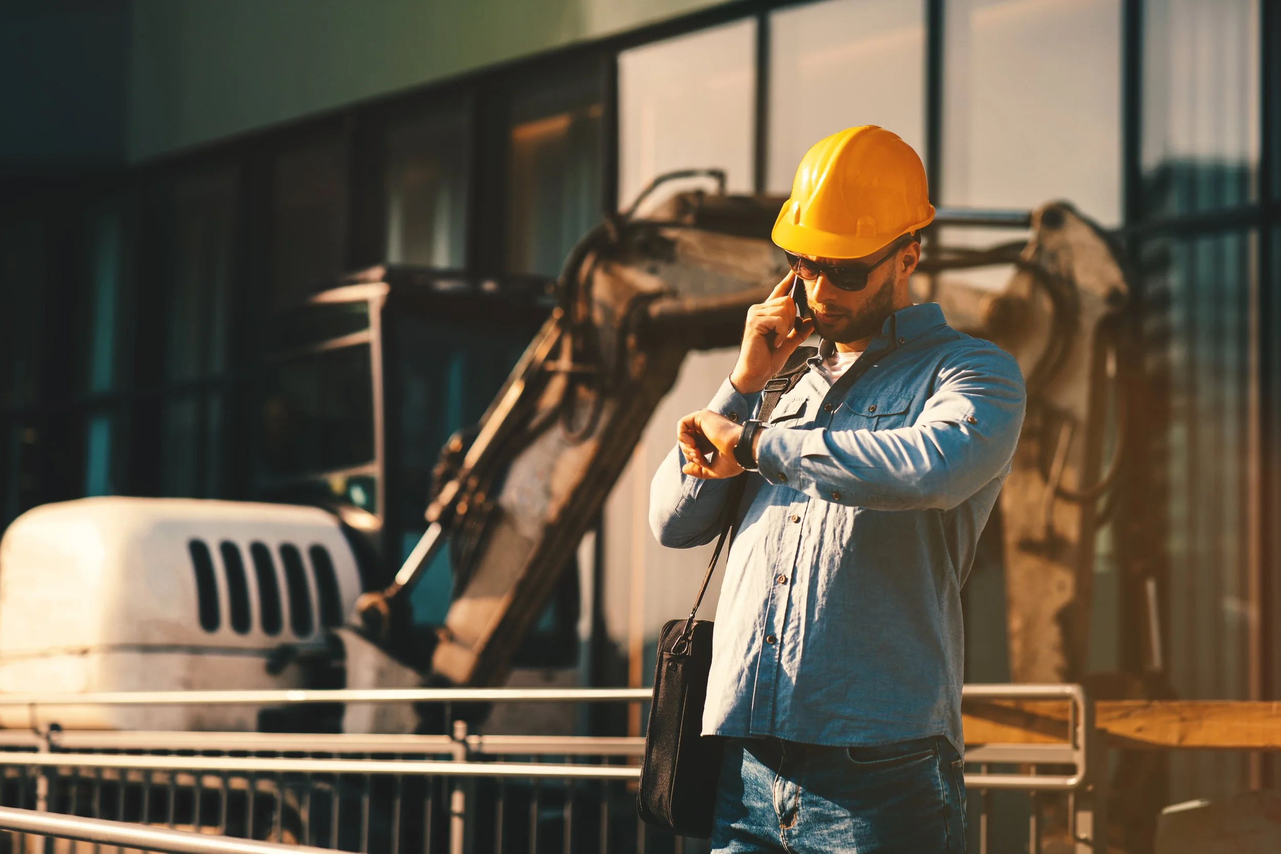 A man wearing a yellow hard hat, sunglasses, a light blue shirt, and jeans, standing outdoors near construction equipment, looking at his wristwatch.