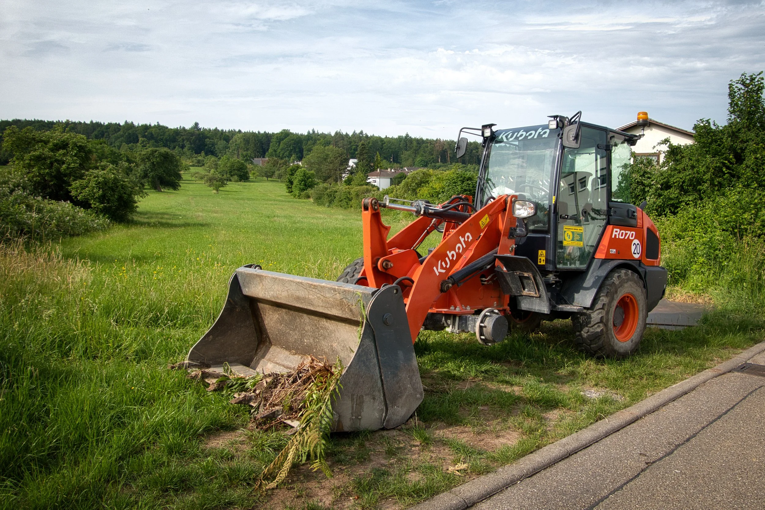 A Kubota backhoe loader parked on a grassy area with trees and houses in the background.