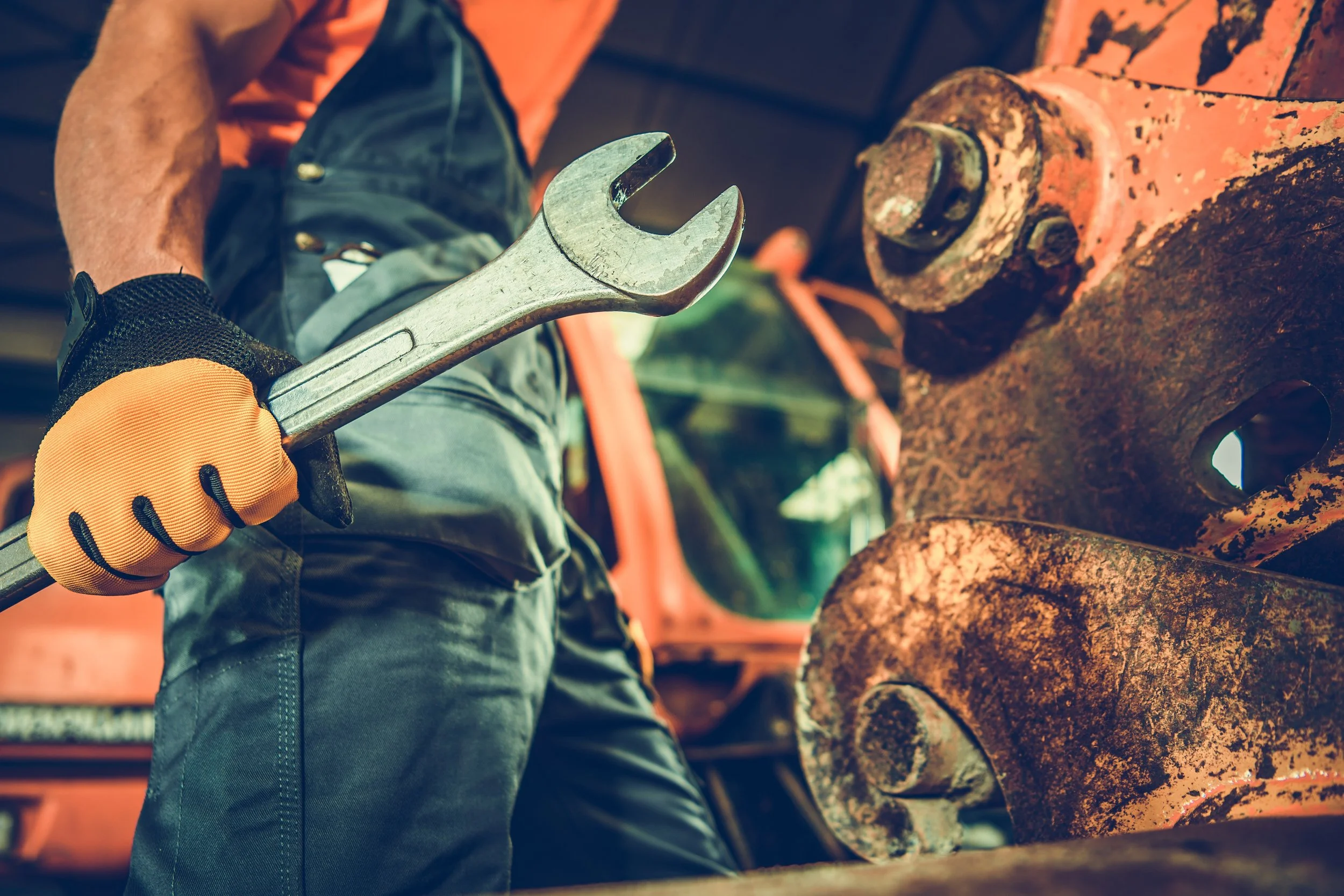 A worker wearing orange gloves and a black watch handles a wrench near a large, rusty pipe with bolts. The worker's clothing includes a black and orange vest and dark pants. The background features additional pipes and industrial equipment.