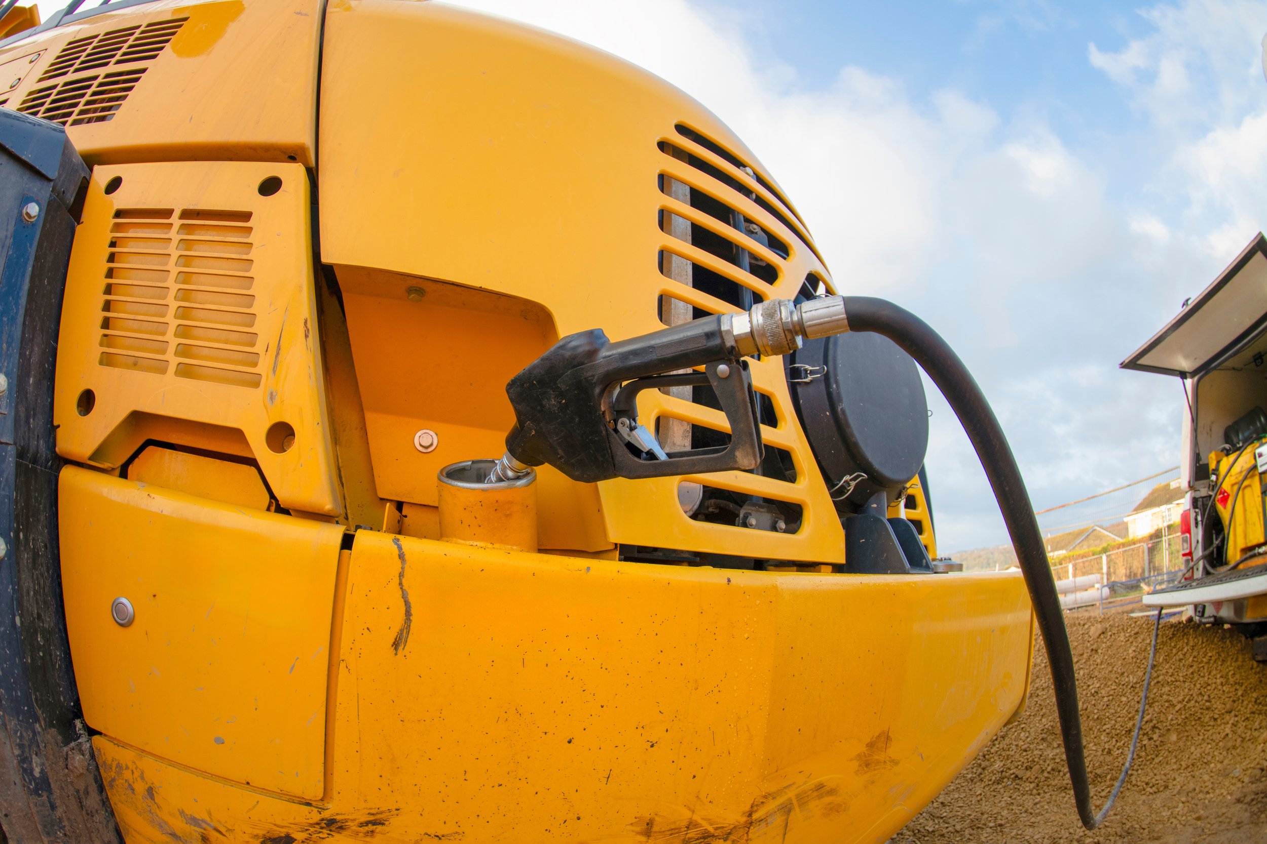 Close-up of a yellow construction vehicle, showing the side panel, air vents, and hydraulic hose connection, against a blue sky.