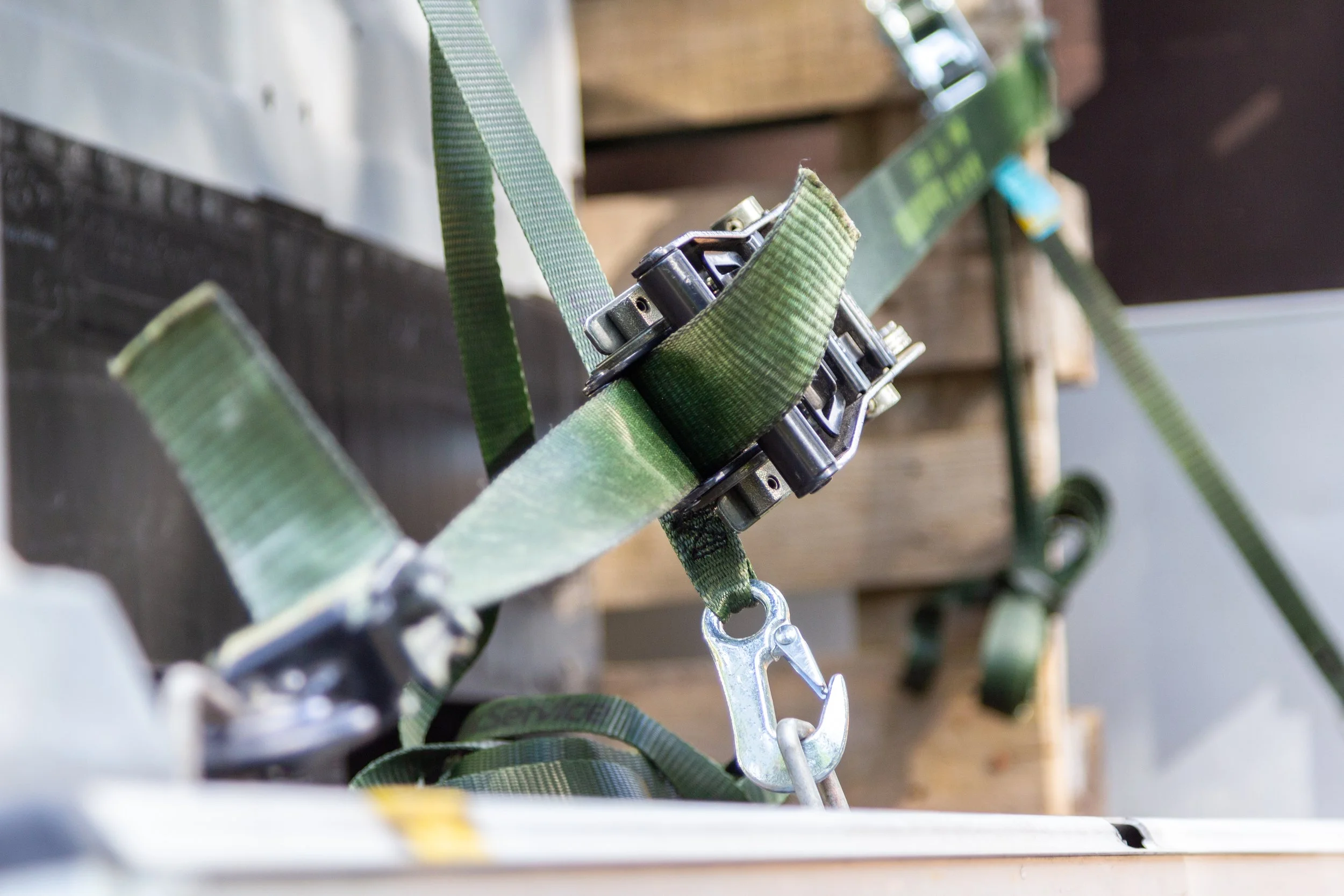 Close-up of a green safety harness with metal hooks and straps, hanging on a wooden structure in an indoor workshop or storage area.