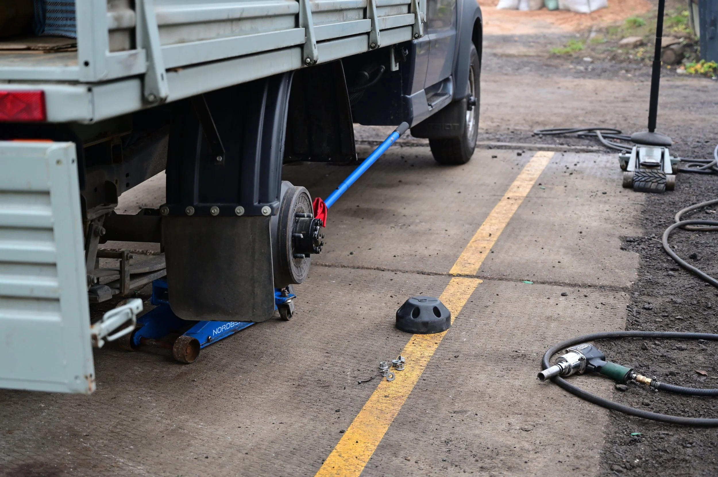 A black truck is being repaired with tools and equipment around it, including a hydraulic jack, hoses, and a black rubber stopper on the ground.