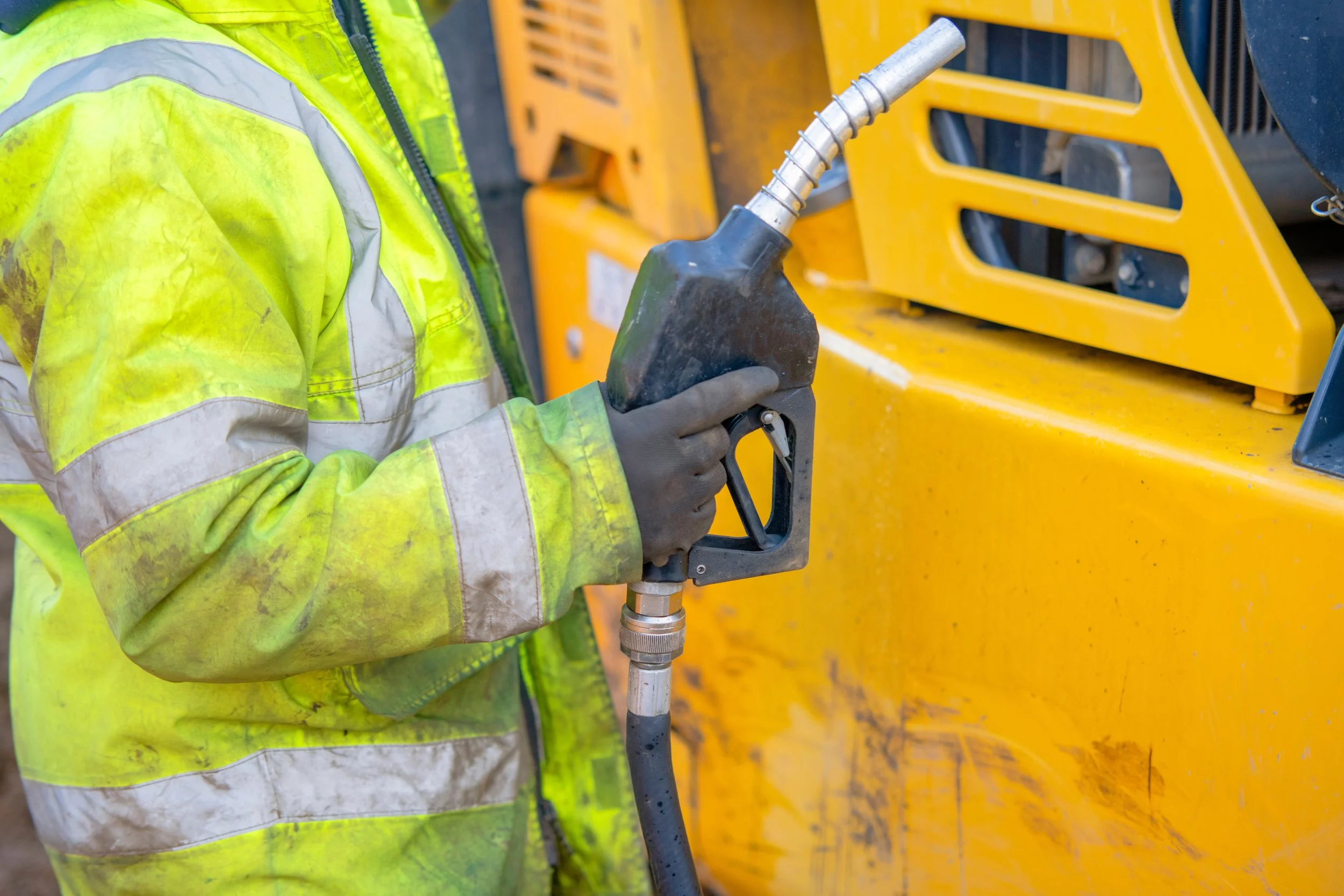 A construction worker in a high-visibility yellow safety jacket holding a tool connected to a yellow machine.