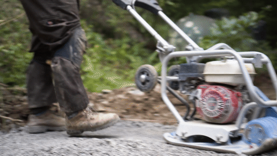 A person operating a plate compactor on a gravel surface outdoors.