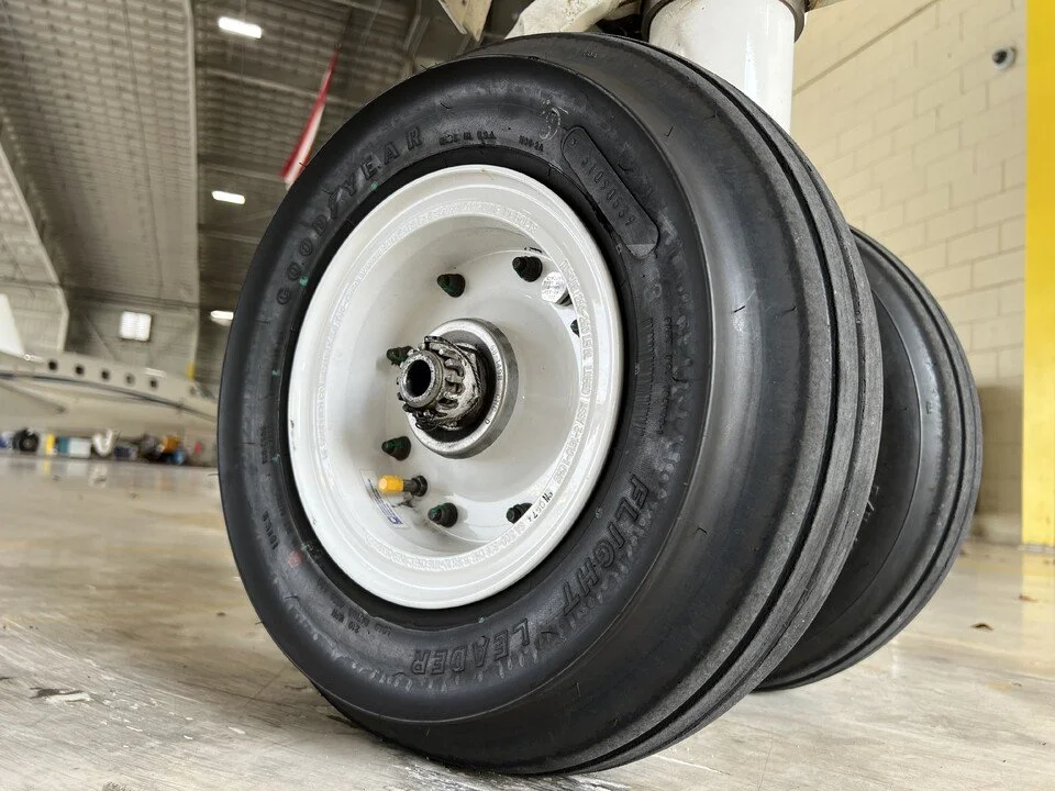 Close-up of a large aircraft wheel with a black tire and white hub, resting on an indoor floor.