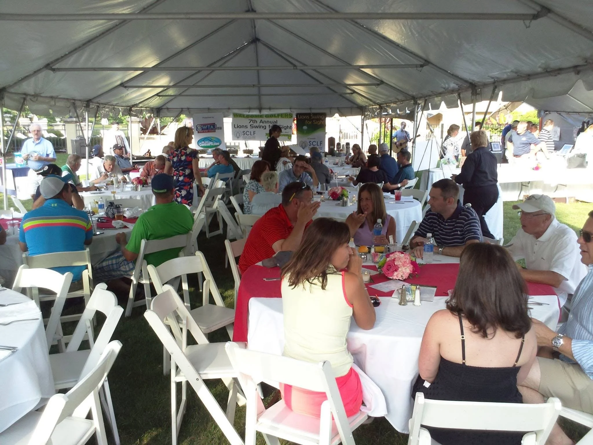 A large outdoor event tent filled with people sitting at round tables with white tablecloths, pink centerpieces, and drinks during a social gathering or festival.