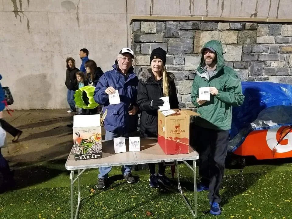 Three people stand behind a table with pamphlets, in front of a stone wall, at night. They appear to be at an event or fundraiser.