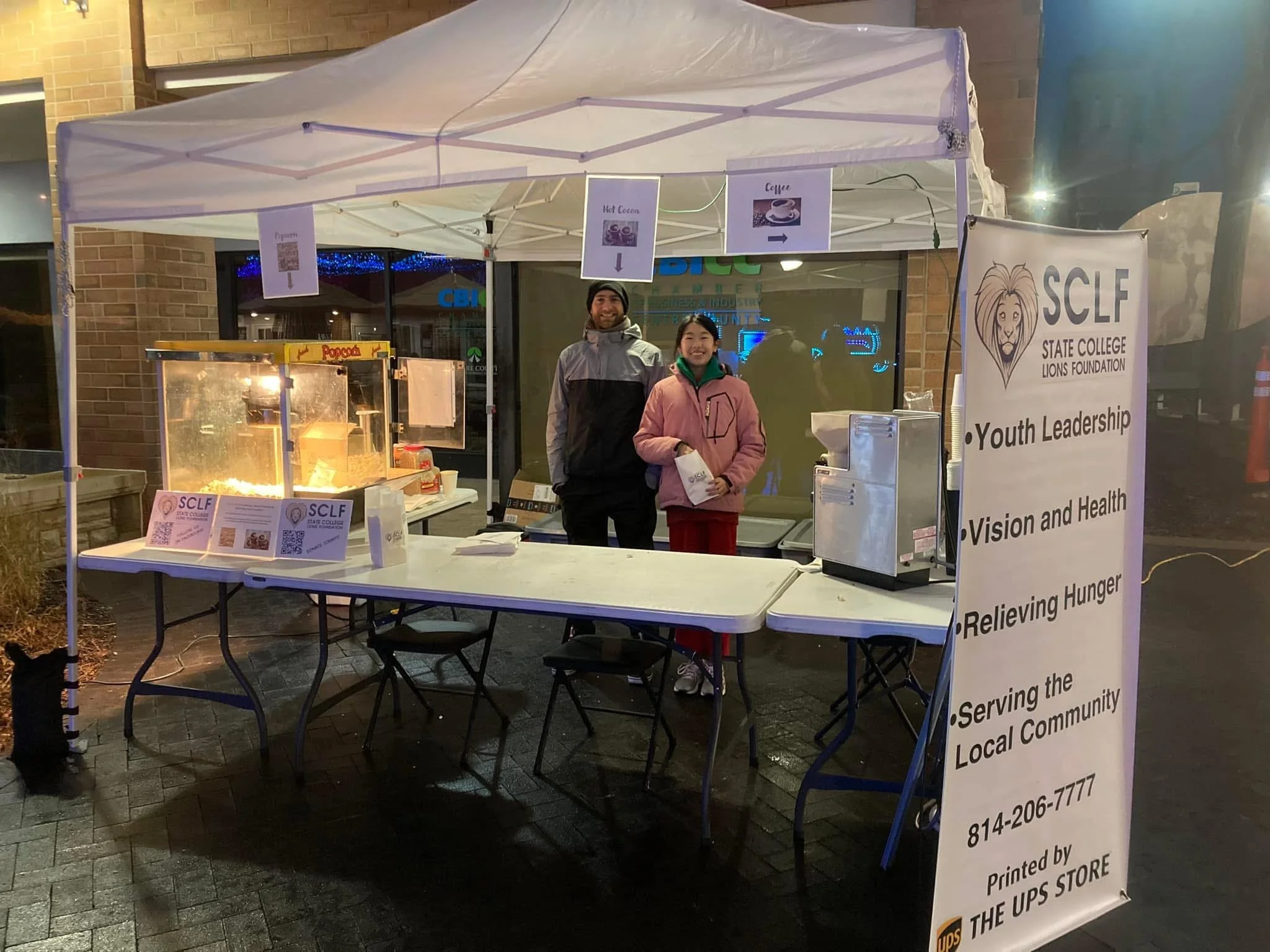 A small outdoor food stand with a white canopy, where two people are smiling at the camera. The stand offers hot popcorn and coffee. A tall white banner displays the Lions Foundation logo and information about serving the local community. There are signs indicating the popcorn and coffee stations, with one pointing to the left and the other to the right. The stand is set up on a paved area outside a brick building, with some decorations in the background.