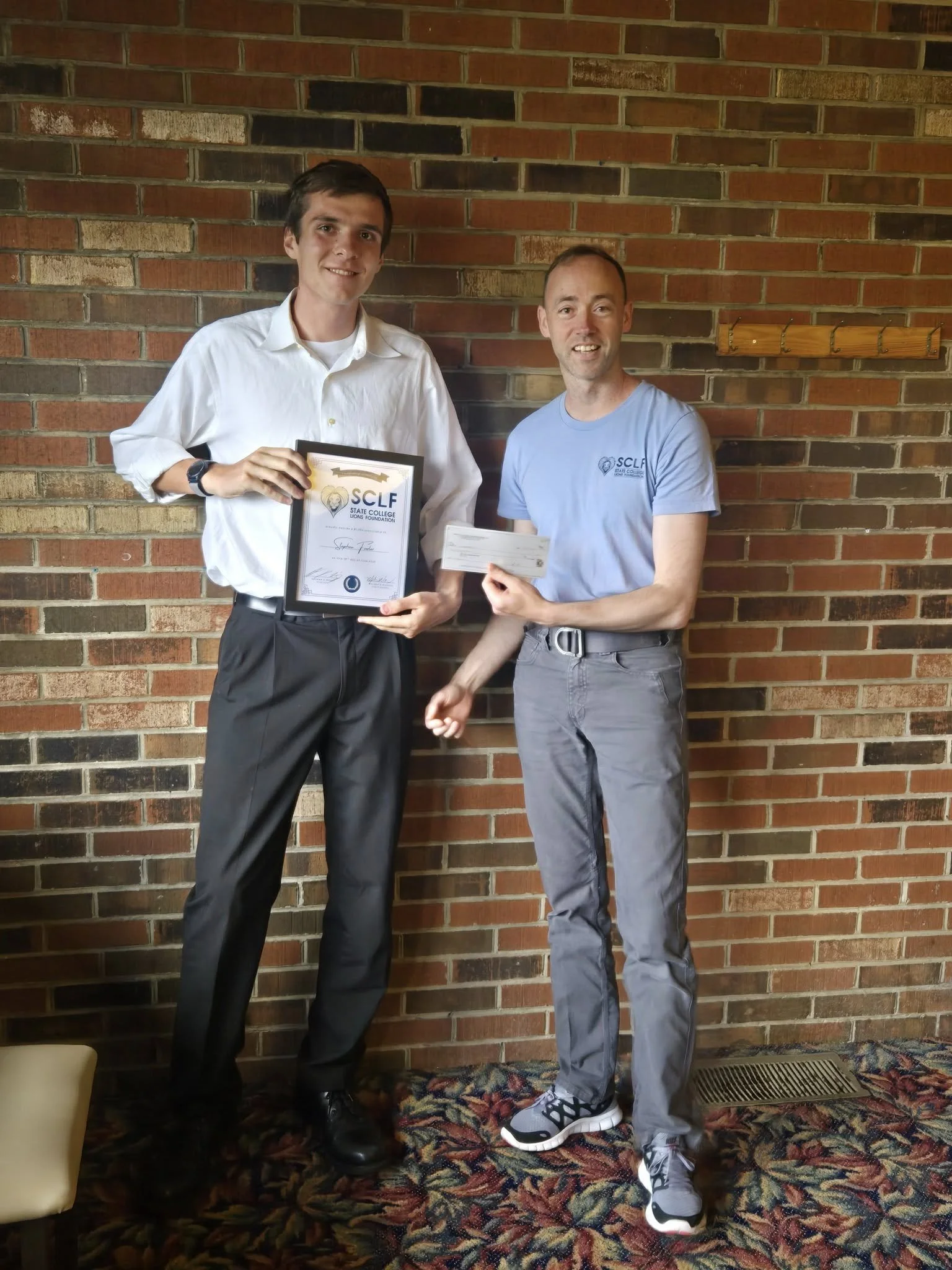 Two men standing in front of a brick wall. One is holding a framed certificate from the State College Lions Foundation. The other is holding a check. The man receiving the award is wearing a white dress shirt and black dress pants. The man presenting the award is wearing a blue t-shirt and gray pants. Both are smiling.