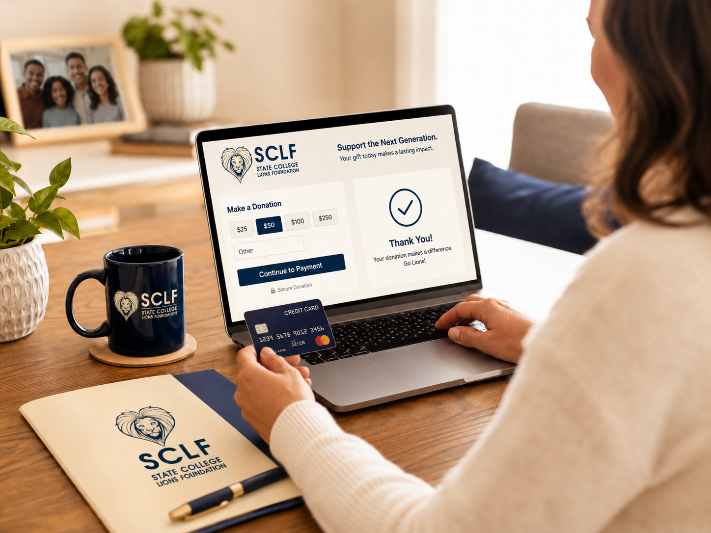 A woman making an online donation for the State College Lions Foundation on a laptop. She holds a credit card and there is a branded coffee mug, a notebook with the foundation logo, a plant, and a framed family photo on a wooden desk.