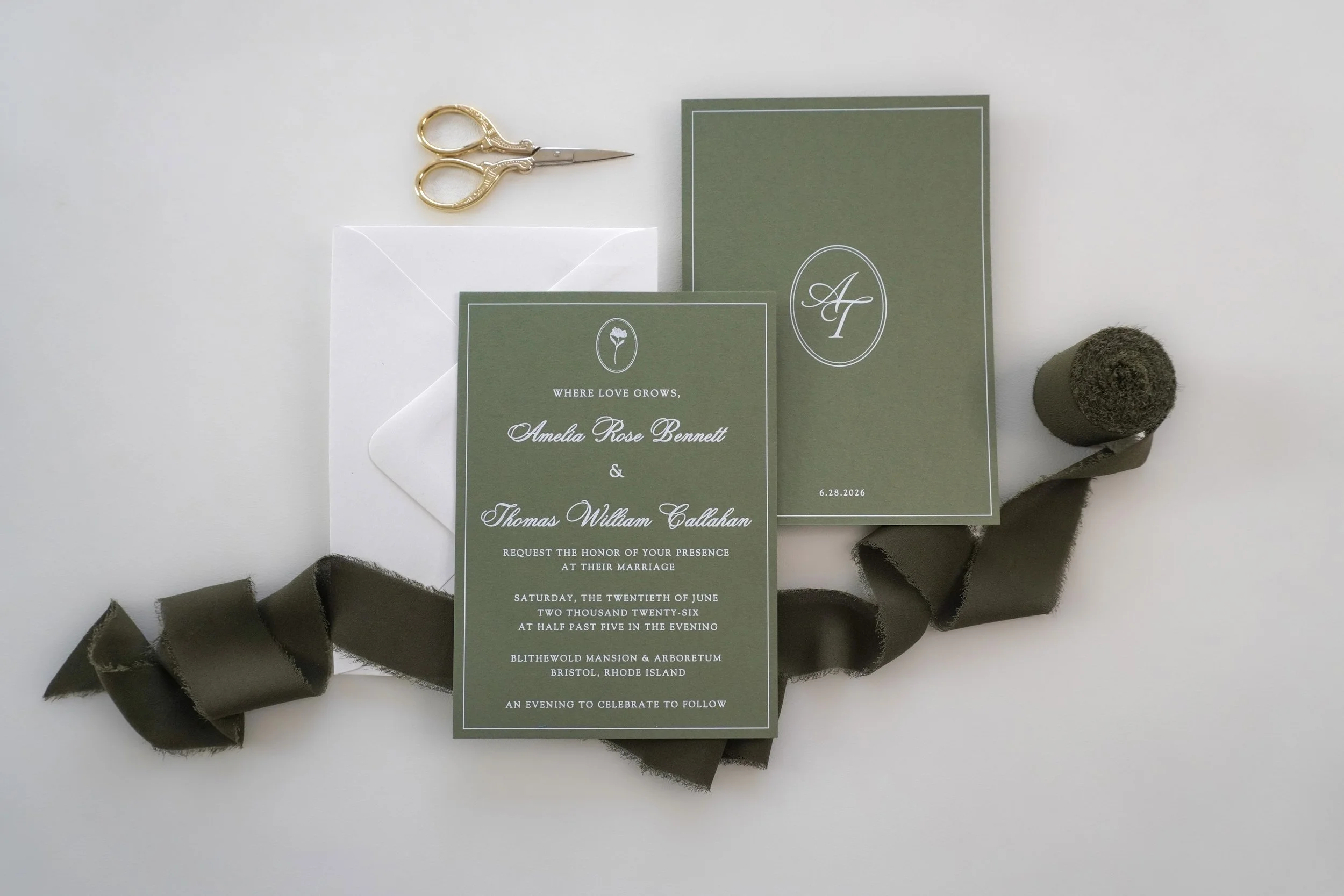 Green wedding invitation card with white text and initials 'A' and 'F', accompanied by a smaller card with a flower icon and the phrase 'Where love grows,' a white envelope, a pair of small scissors with gold handles, a roll of green ribbon, and dark green ribbon curled around the items, all arranged on a white surface.