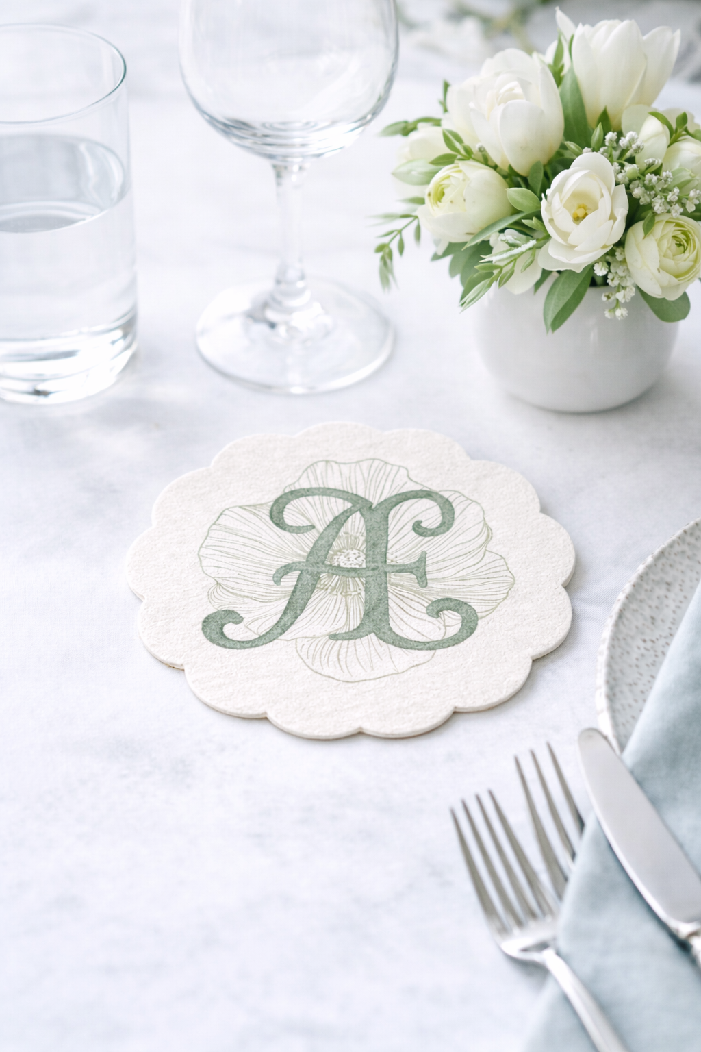 Elegant table setting with a flower arrangement in a white vase, a glass of water, empty wine glass, silverware, and a decorative napkin with a monogram letter 'A' on a scalloped edge.