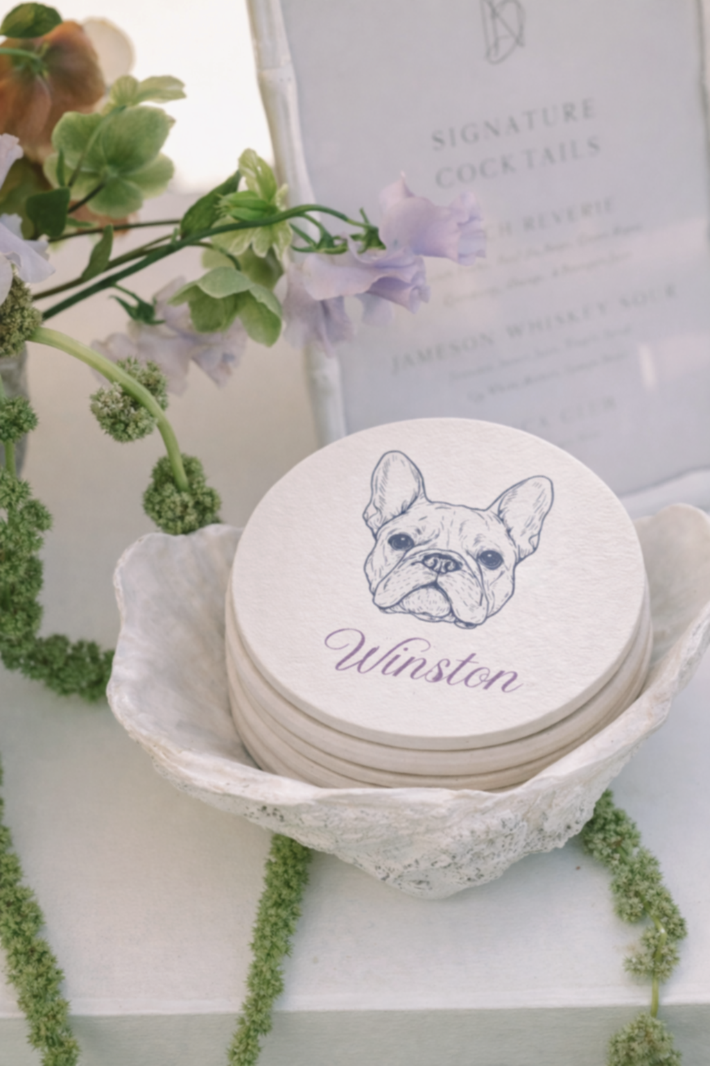 Stack of circular cards with a French Bulldog illustration and the name Winston, placed in a shell-shaped dish; surrounded by white and green floral decorations and an out-of-focus sign in the background.