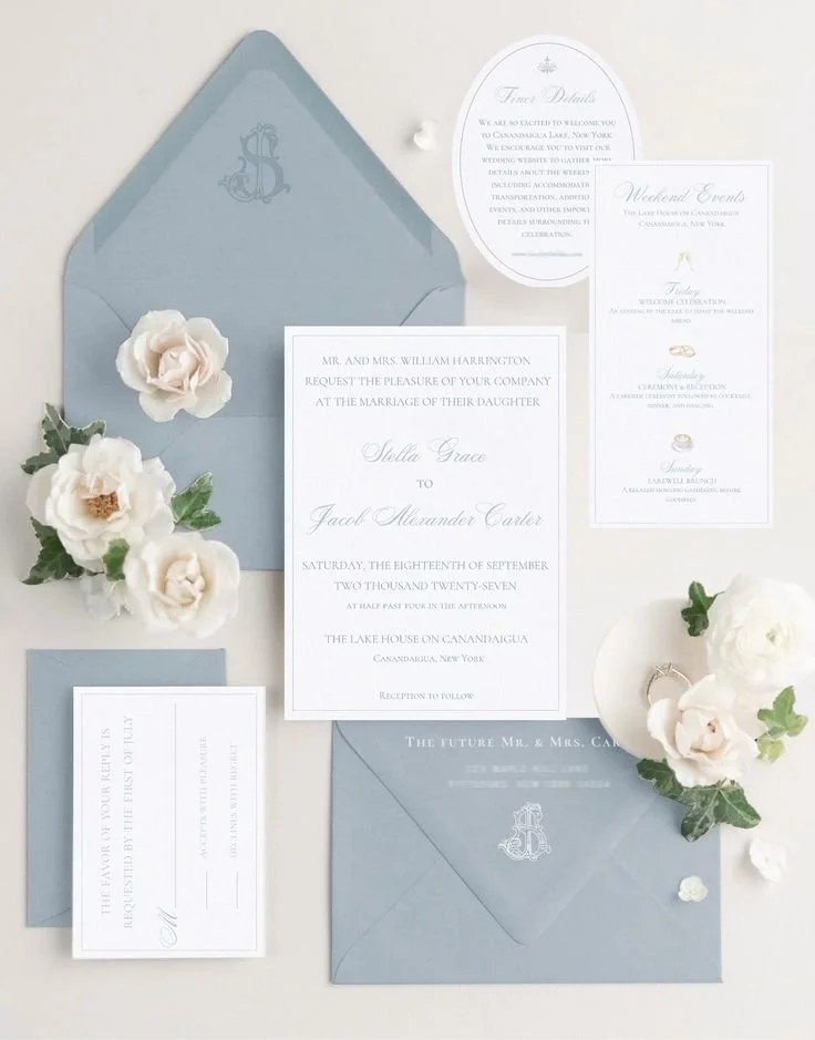 Elegant wedding invitations in white and light blue, adorned with white flowers and greenery, layered with RSVP and event details.