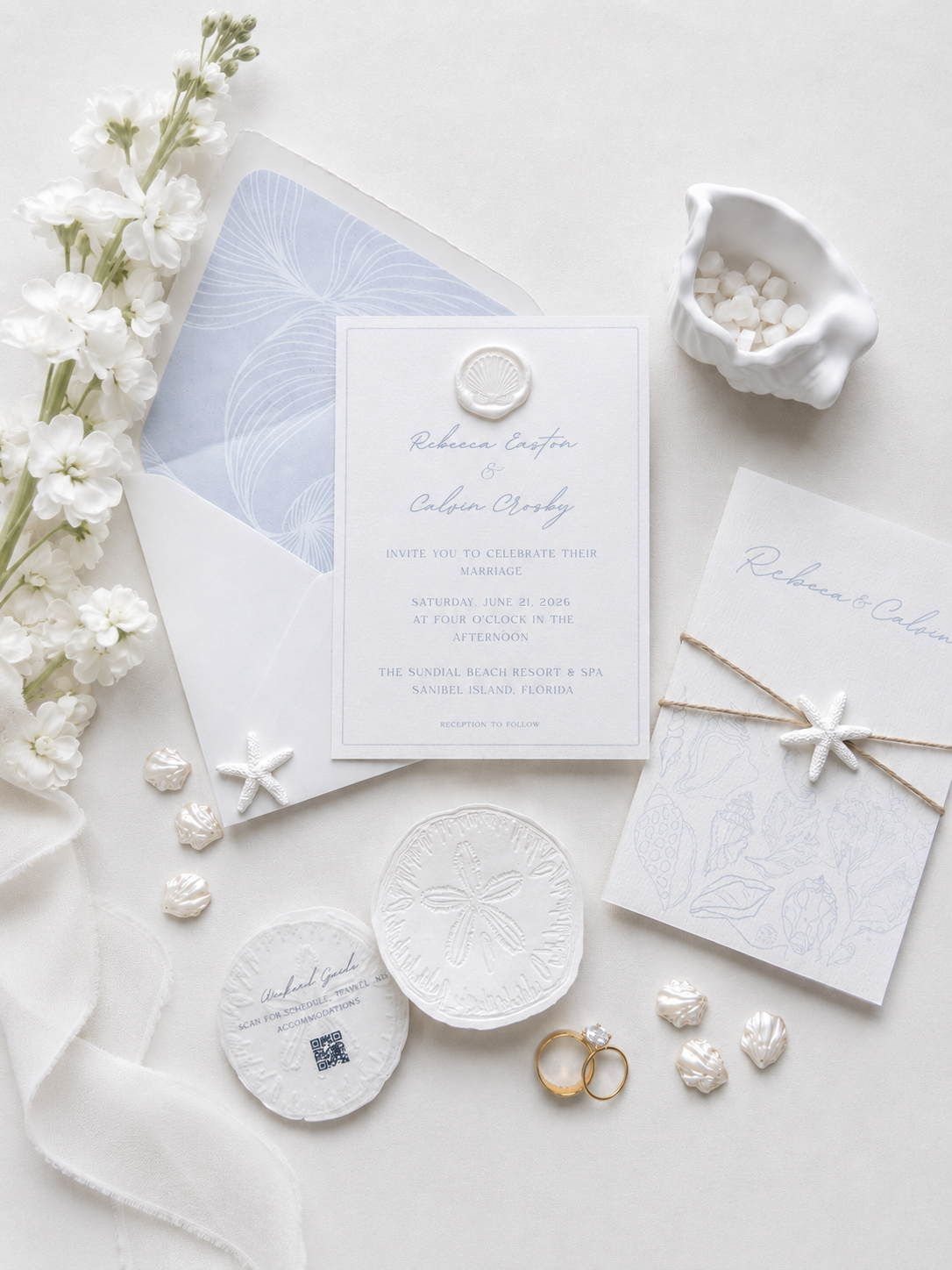 Beautiful wedding invitation suite with white and blue decor, seashell and starfish motifs, gold wedding bands, an envelope with a blue liner, and white floral accents.