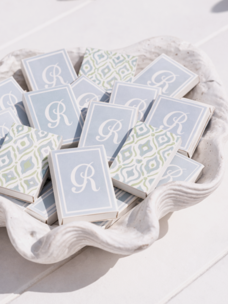 A white, decorative shell-shaped bowl filled with small matchboxes featuring a blue background and white letter 'R' with some patterned matchboxes in green, blue, and white, on a white surface.