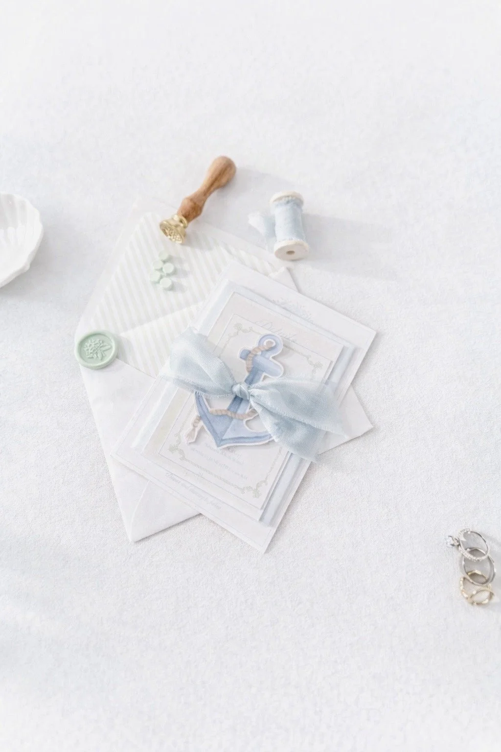 A collection of wedding or baby shower items including a card with an anchor, a gift envelope, a ribbon tied around the card, a wax seal stamp, spools of light-colored thread, and small decorative items arranged on a white textured surface.