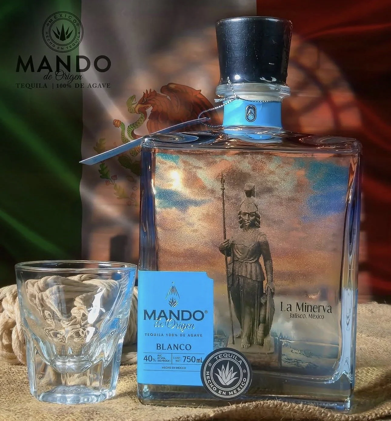 A clear glass bottle of Mando de Origen tequila with a label showing a woman holding a speared staff, along with a blue label indicating it is a Blanco variety with 40% alcohol content and 750 mL volume. A small, empty glass sits in front of the bottle. Behind, a partially visible bag of Mexican Mando de Origen tequila with a logo and illustration of a lion and snake, and a Mexican flag in the background.