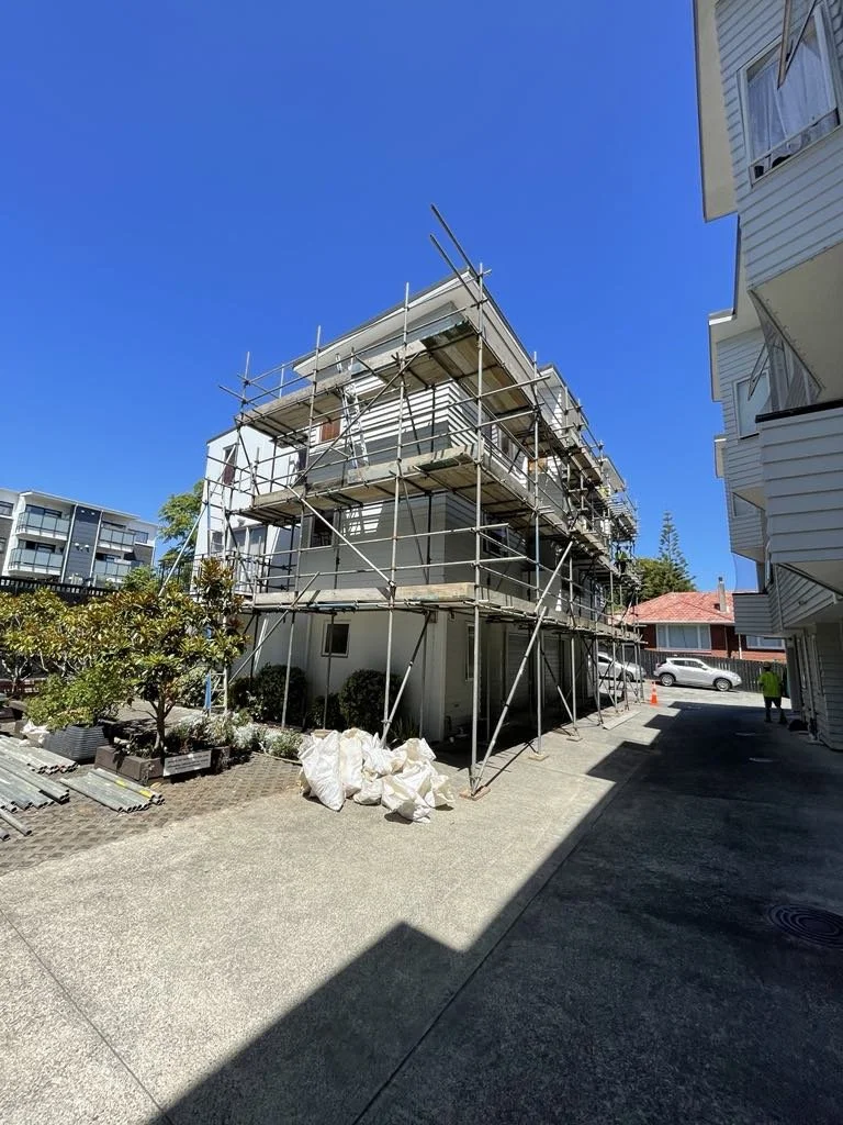 Building under construction with scaffolding on a sunny day, residential area with neighboring apartment buildings, trees, and parked cars.