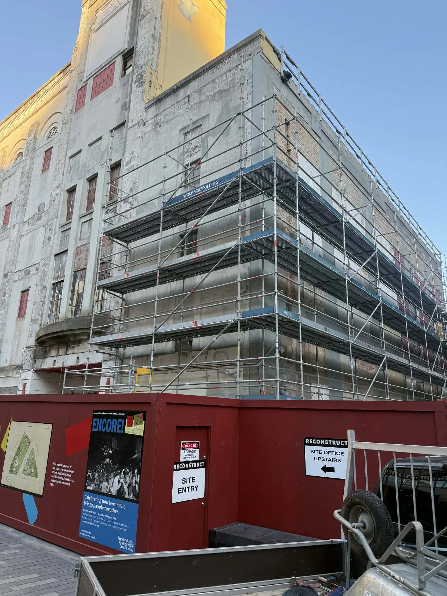 A multi-story building under renovation with scaffolding surrounding it. There are signs on a red construction fence indicating site entry and directions, along with posters about local events and safety notices.
