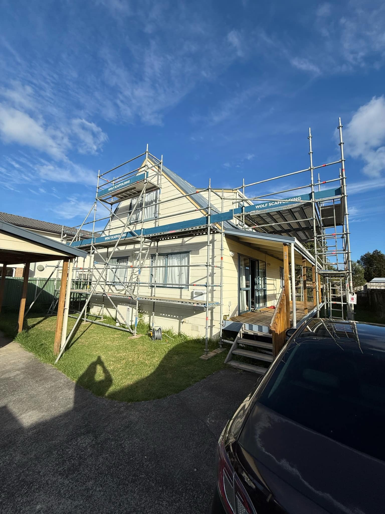 House with scaffolding around the roof under a partly cloudy blue sky, a black car parked in the foreground, and a small patch of grass and driveway in the yard.