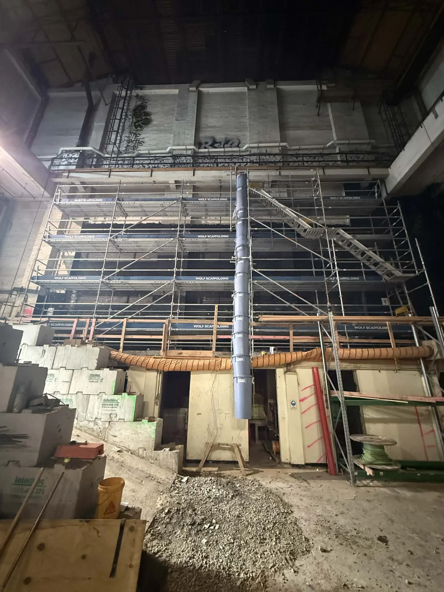 Interior of a construction site with scaffolding, unfinished walls, construction materials, and debris on the ground.