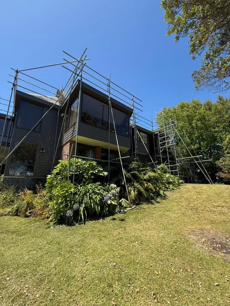 Scaffolding around a modern house with large windows, surrounded by green plants and trees under a clear blue sky.