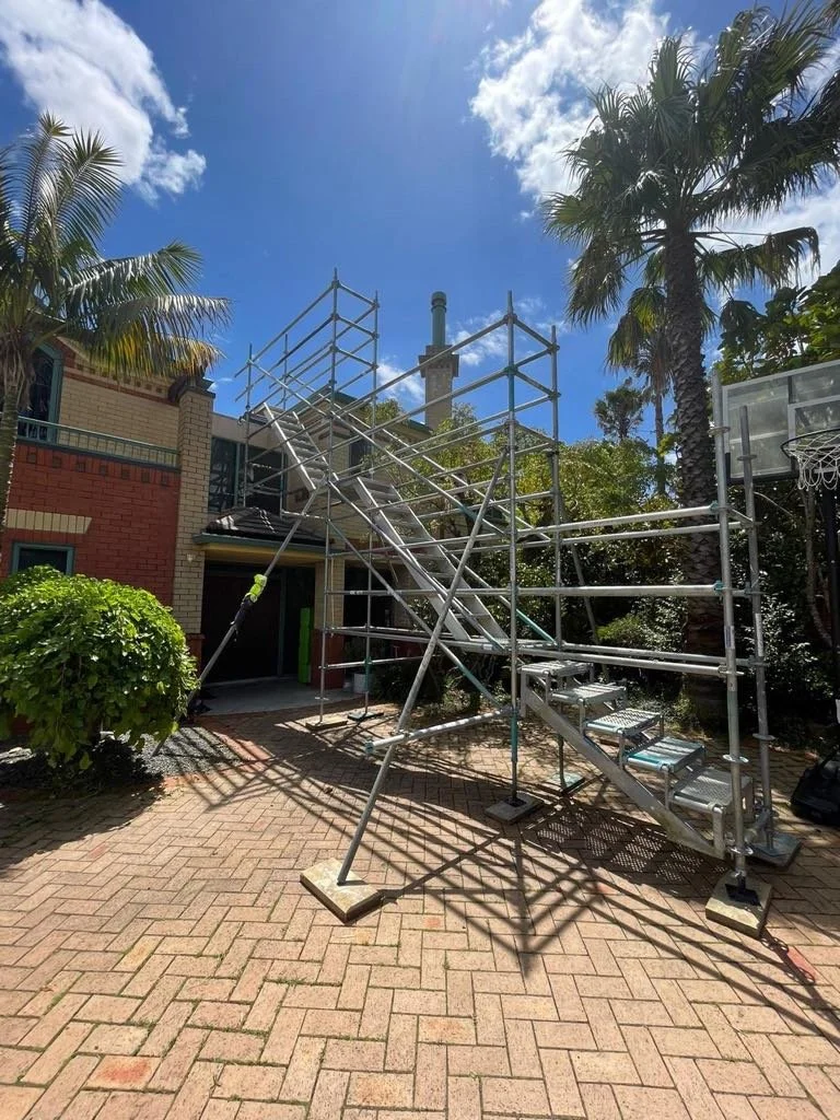 Scaffolding set up on a brick patio in front of a house with palm trees in the background, a basketball hoop on the right, and a partly cloudy sky overhead.