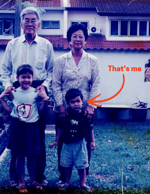 A family photo with an elderly couple and two children standing outdoors in front of a house with a red tiled roof. The elderly man has gray hair and glasses, wearing a white shirt. The elderly woman has short black hair, wearing a patterned blouse. A young girl stands on the left, wearing a white t-shirt with a cartoon character and a dark skirt, holding her shirt. A young boy stands on the right, wearing a black t-shirt and gray shorts, with a caption pointing to him that says, 'That's me'.