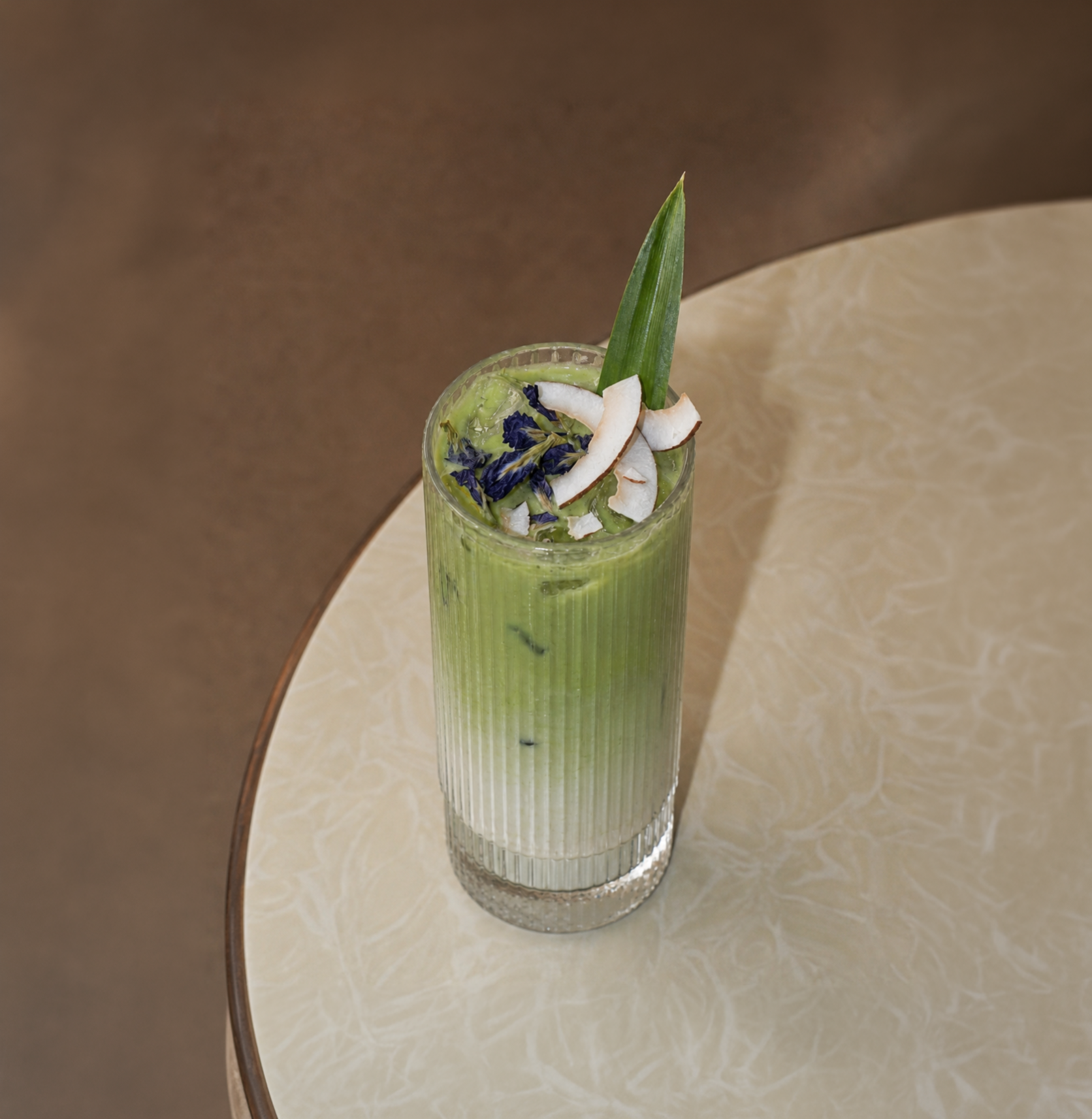 A tall, clear glass filled with a bright green smoothie, topped with coconut flakes, blue edible flowers, and a green leaf, placed on a beige marble table.