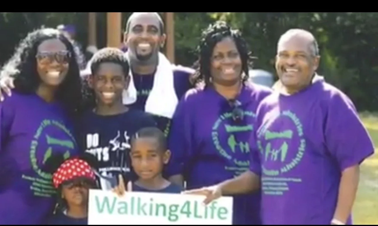 Group of diverse people wearing purple Walking4Life t-shirts, smiling outdoors, holding a sign that says Walking4Life.