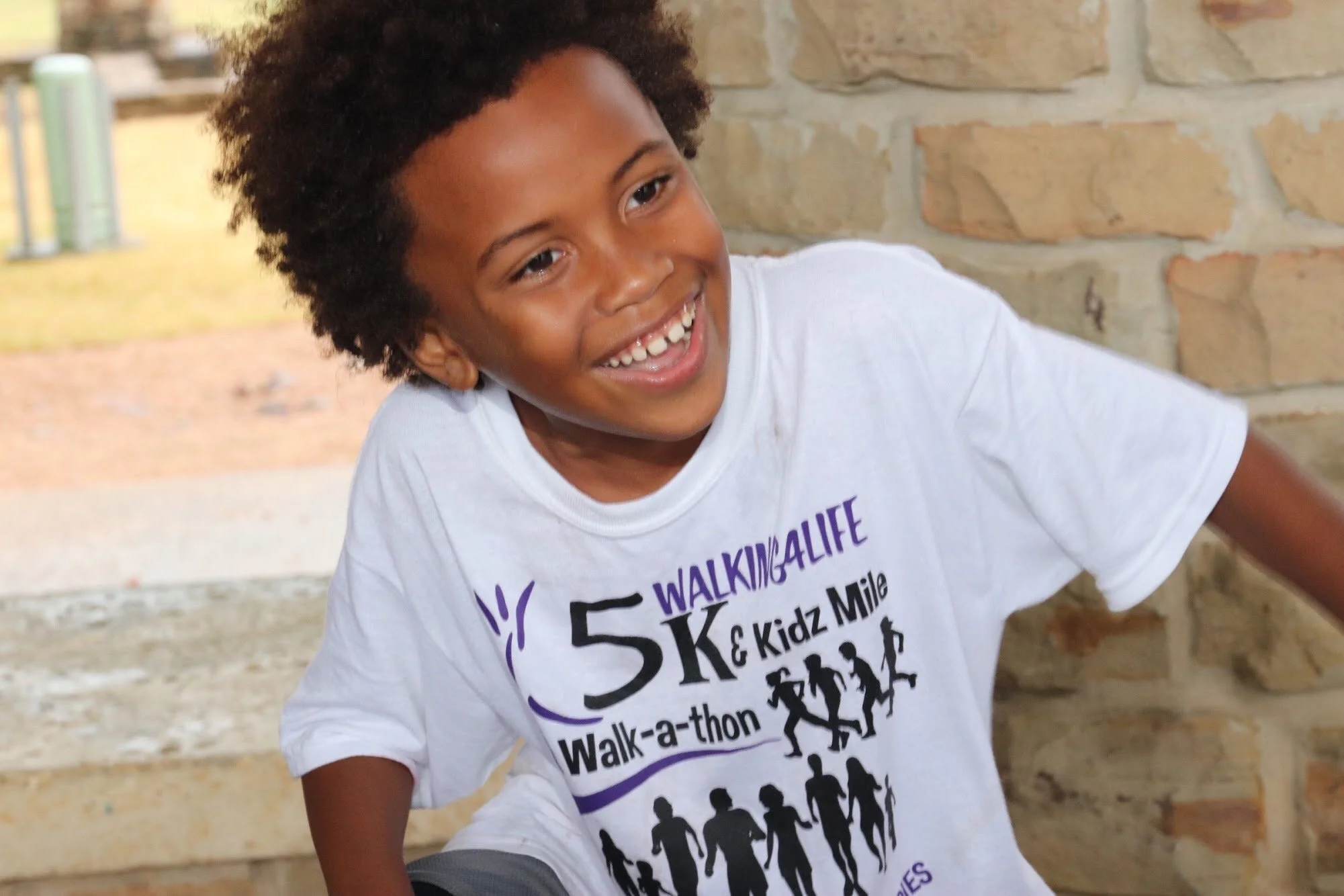 Young person with curly hair smiling, wearing a white event T-shirt with purple and black print, outdoors near a brick wall.